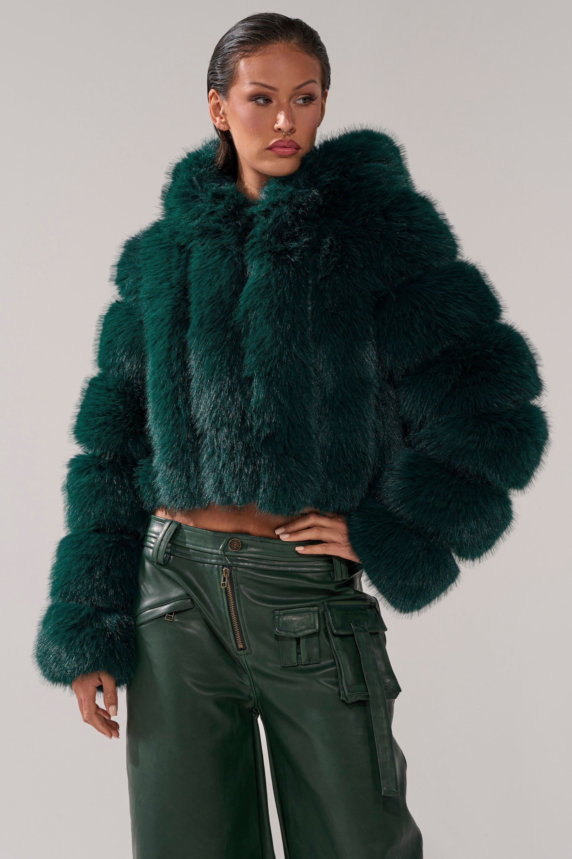 A woman stands against a plain light background wearing the JOLENE FAUX FUR COAT IN EMERALD GREEN, featuring a hood, paired with matching high-waisted leather pants with multiple pockets. 0