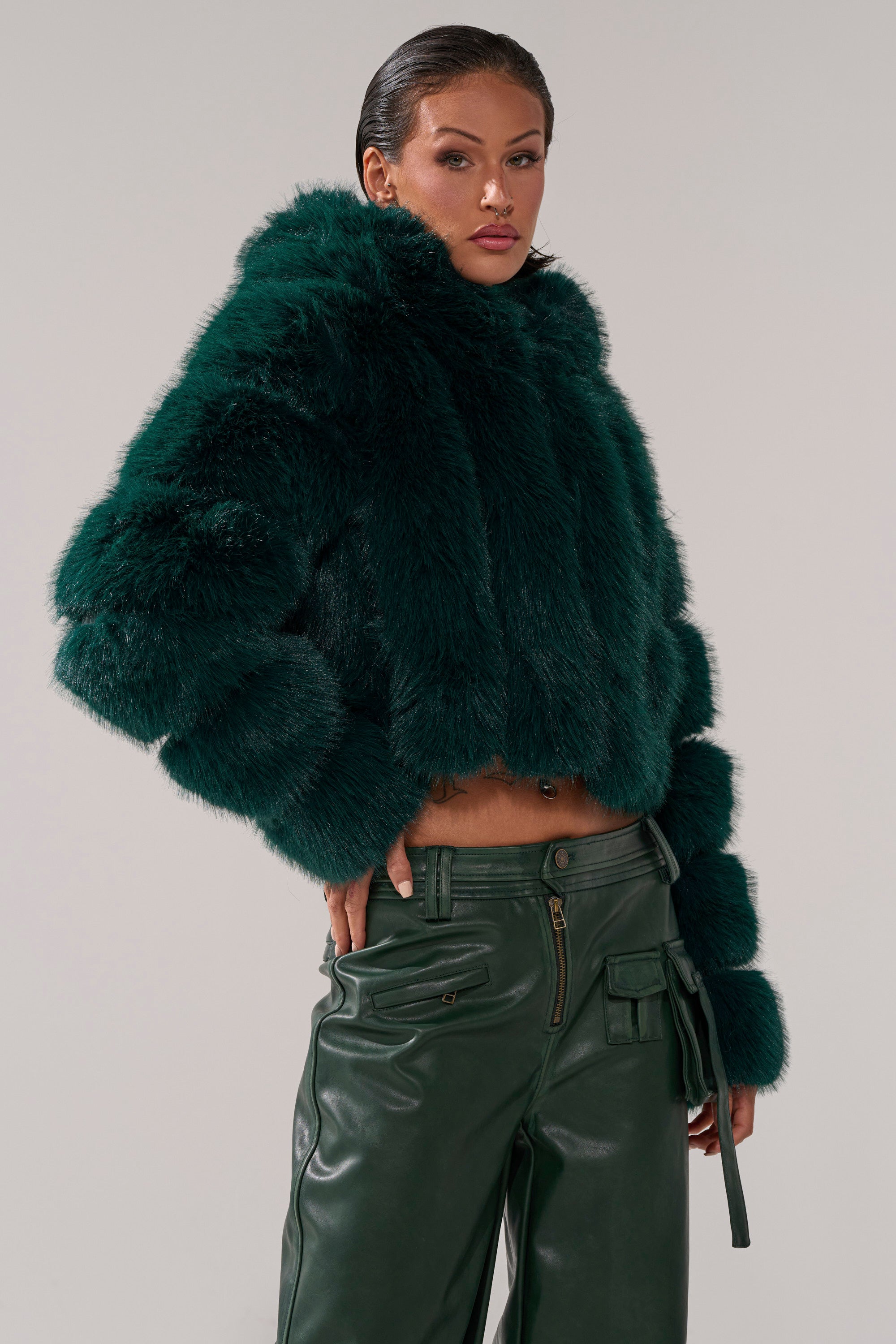 Wearing the JOLENE FAUX FUR COAT IN EMERALD GREEN, a person poses with one hand in their pocket against a plain light background, pairing the fluffy hooded coat with matching green leather pants. 3