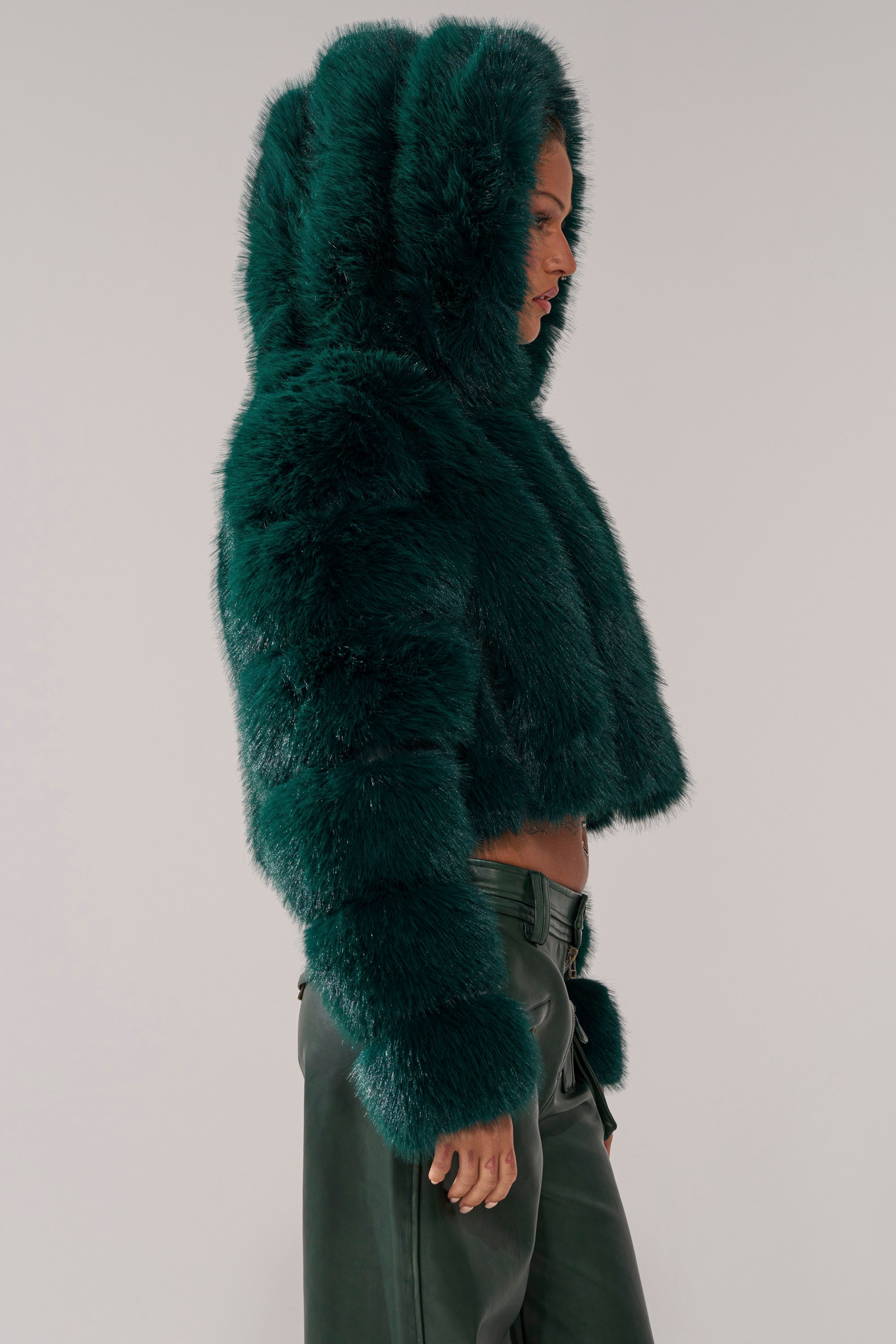 A person stands in profile wearing the JOLENE FAUX FUR COAT IN EMERALD GREEN, paired with matching pants against a plain light background. 4