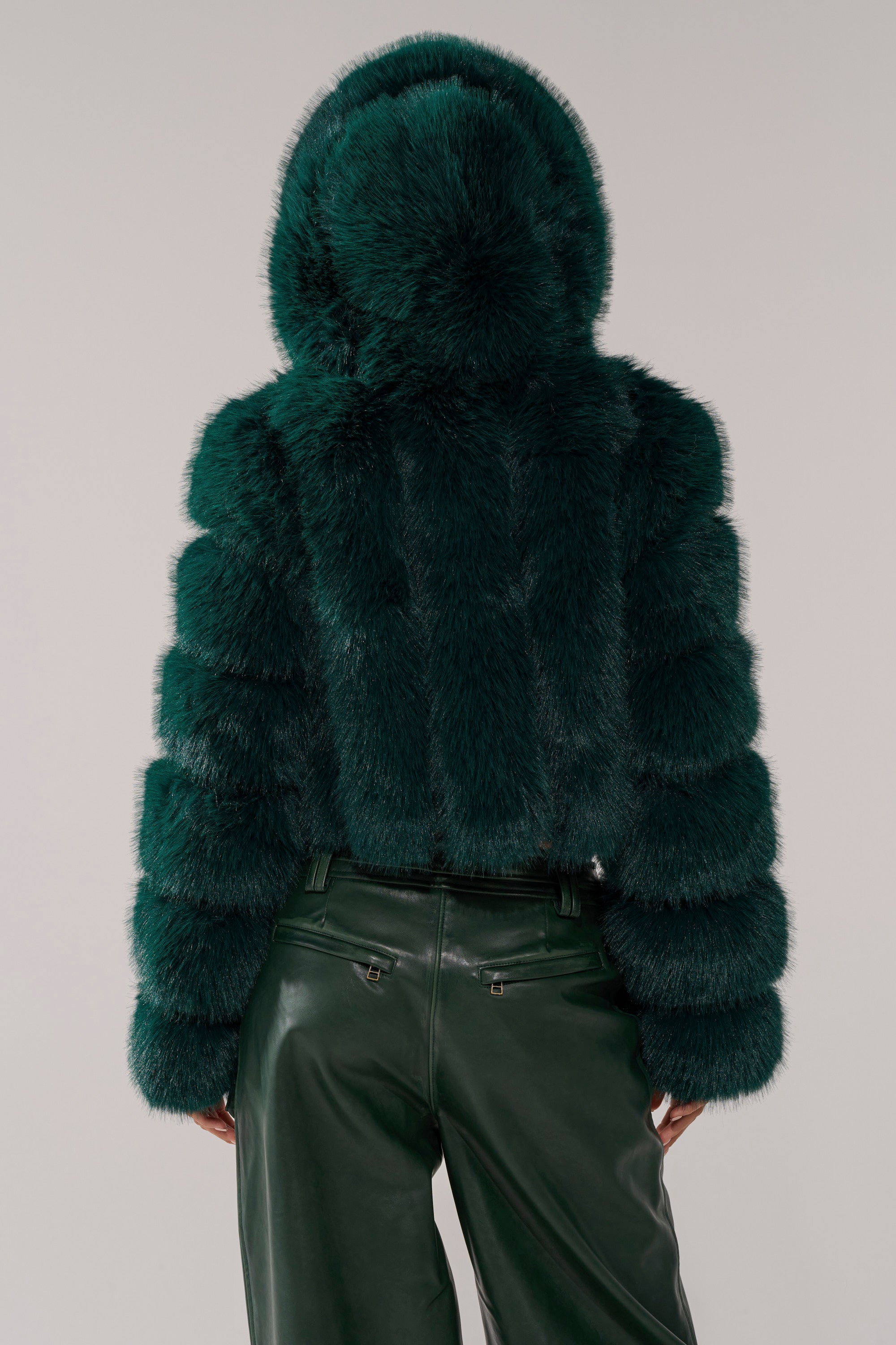 A person is shown from the back against a light gray background wearing the JOLENE FAUX FUR COAT IN EMERALD GREEN with a hood, paired with high-waisted, dark green leather pants. 5