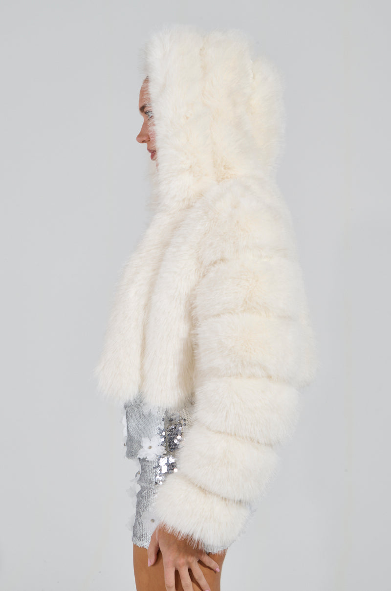Wearing the JOLENE FAUX FUR COAT IN WHITE, a person poses in profile, showcasing a silver sequined outfit. The plain light-colored background highlights the textured fur coat and shimmering ensemble, creating a striking silhouette.