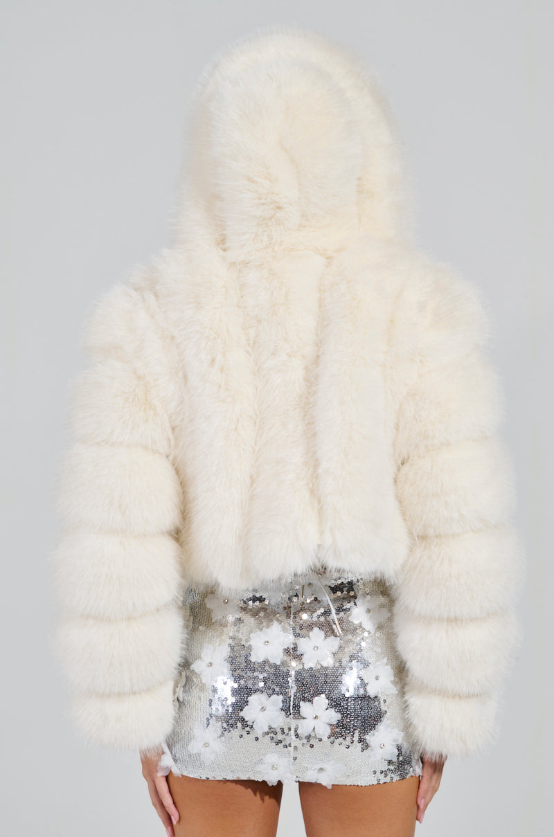 A person stands with their back to the camera, wrapped in a fluffy, hooded JOLENE Faux Fur Coat in white and a silver sequin skirt adorned with floral patterns.
