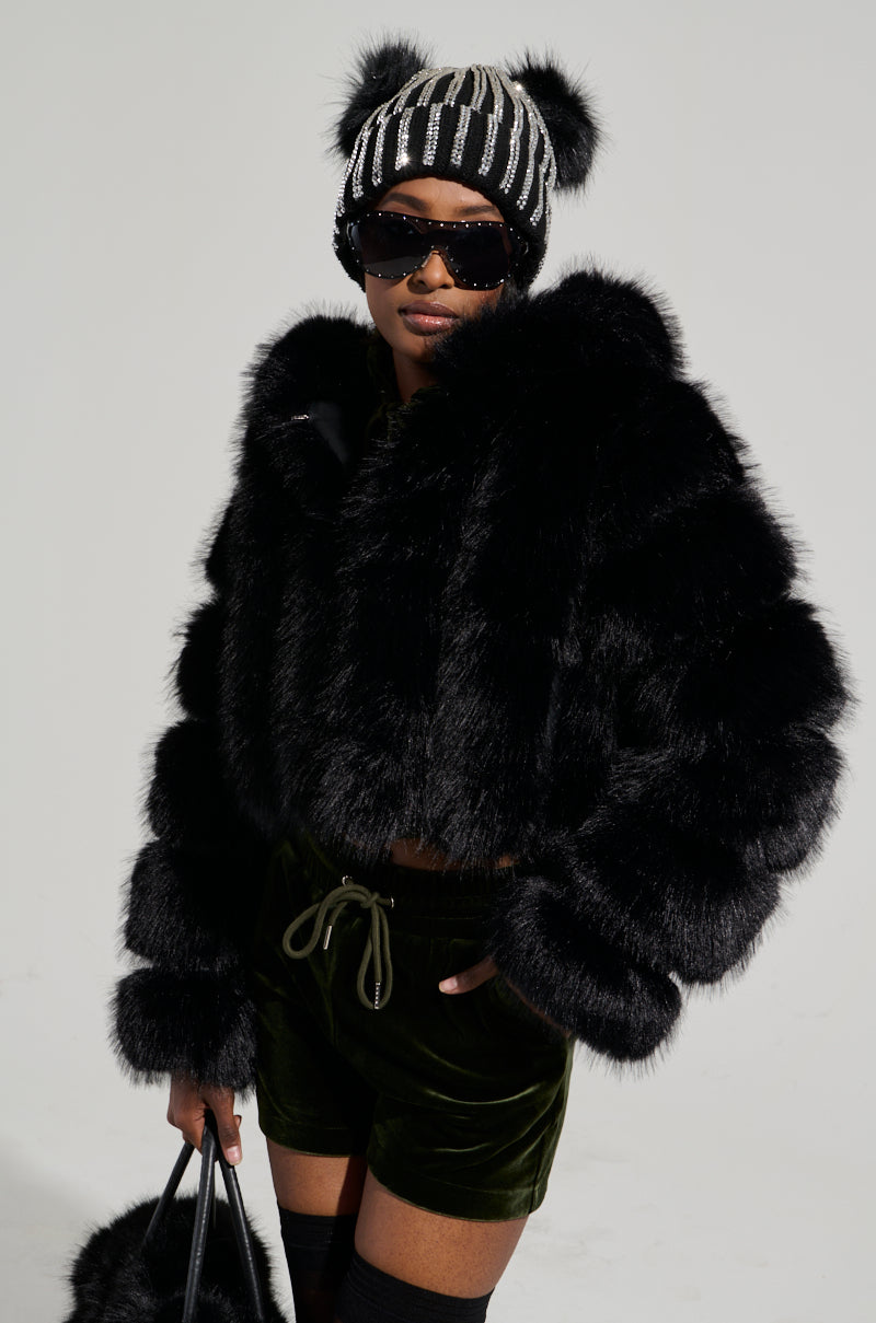 Wearing the JOLENE FAUX FUR COAT IN BLACK, a person stylishly pairs it with dark green velvet shorts and a playful black and white beanie adorned with pom-poms. They accessorize their look with sunglasses and hold a black bag, posing elegantly against a light gray backdrop.
