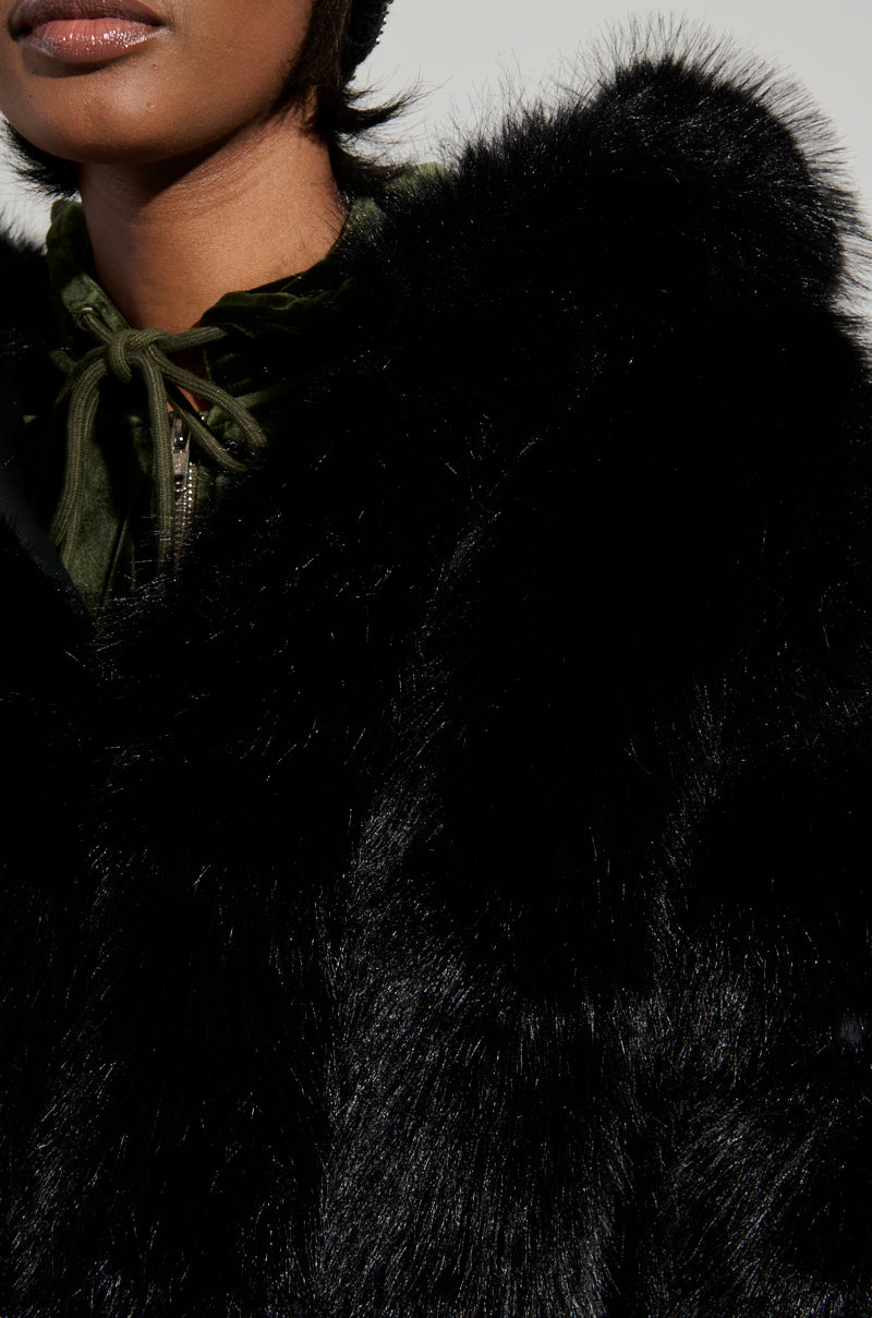 A person wearing the JOLENE FAUX FUR COAT IN BLACK, featuring a stylish sherpa-lined hood and a touch of green fabric accentuating the collar. The emphasis is on the luxurious texture of the faux fur and the striking contrast with the green detail, while the background remains softly blurred.