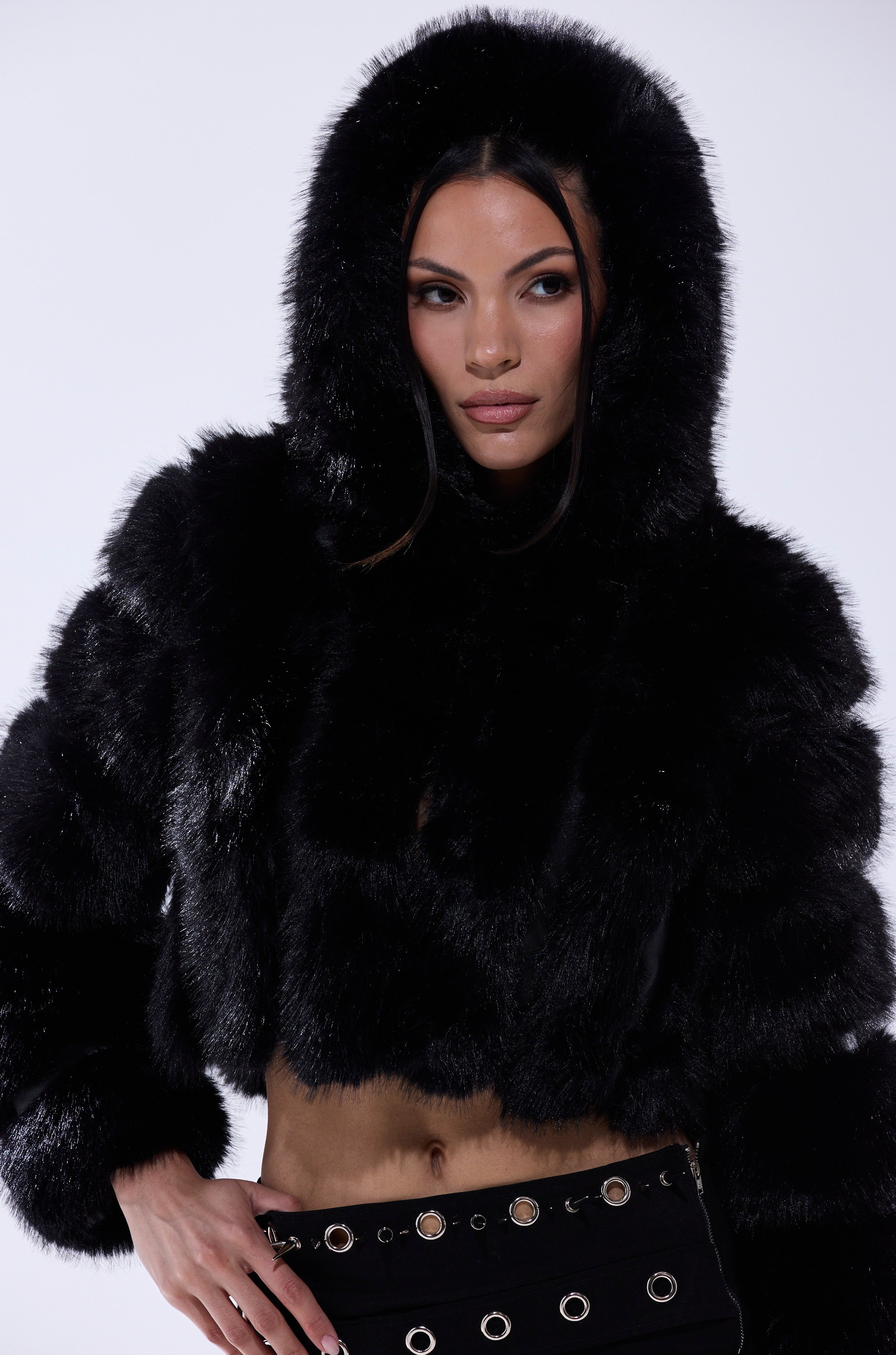 A woman with long dark hair wears the JOLENE FAUX FUR COAT IN BLACK, featuring a sherpa-lined hood, paired with black pants with metal grommets, standing against a plain light background.