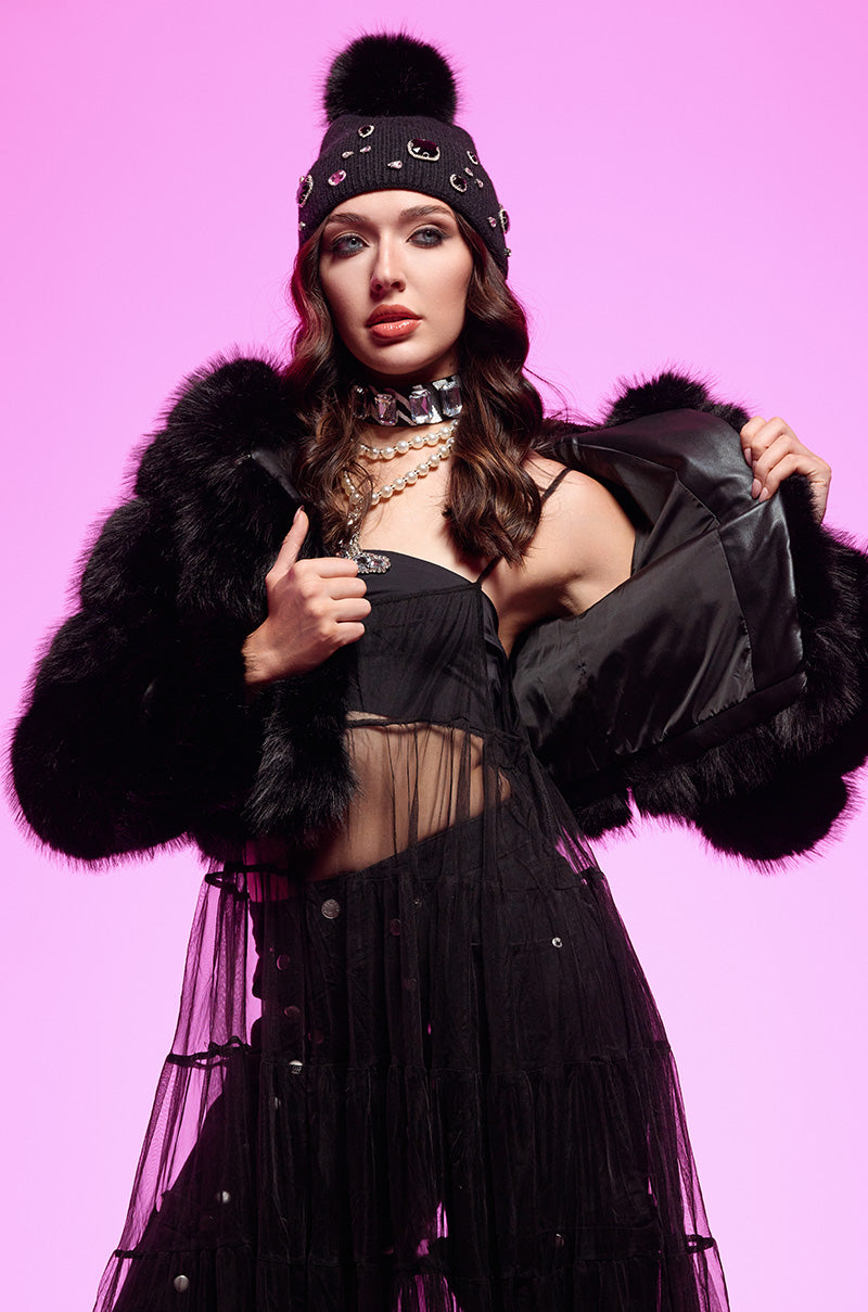 A woman poses against a pink background, showcasing the JOLENE FAUX FUR COAT IN BLACK layered over a sheer black dress and accessorized with a beanie featuring metal embellishments. She holds the sherpa-lined hood open, revealing a strapless top underneath along with layered necklaces.