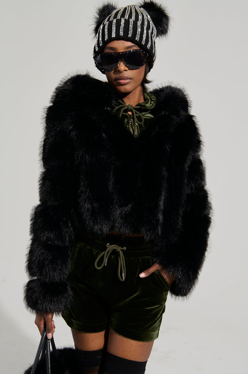 Person wearing the Jolene Faux Fur Coat in Black, paired with a black knit hat featuring pom-poms, sunglasses, and green velvet shorts. They hold a black bag and wear knee-high black socks against a plain background.