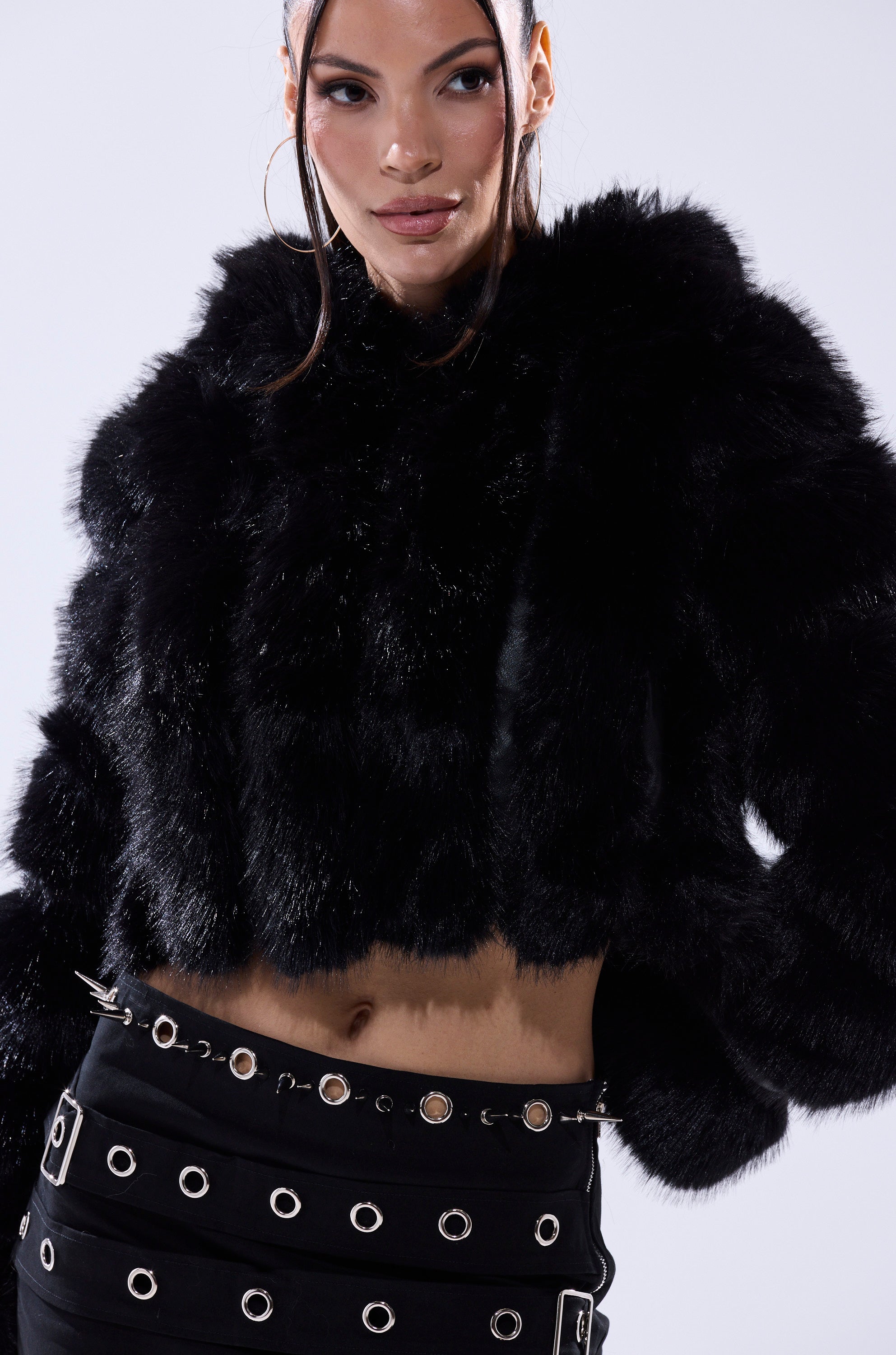 A woman wears the JOLENE FAUX FUR COAT IN BLACK and a black skirt with silver grommets and studs, posing against a light background.