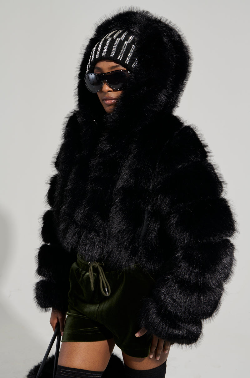 Wearing the JOLENE FAUX FUR COAT IN BLACK, a person showcases their chic style with stylish black sunglasses, a striped beanie, and green velvet drawstring shorts. They carry a sleek black bag while standing against a simple backdrop.