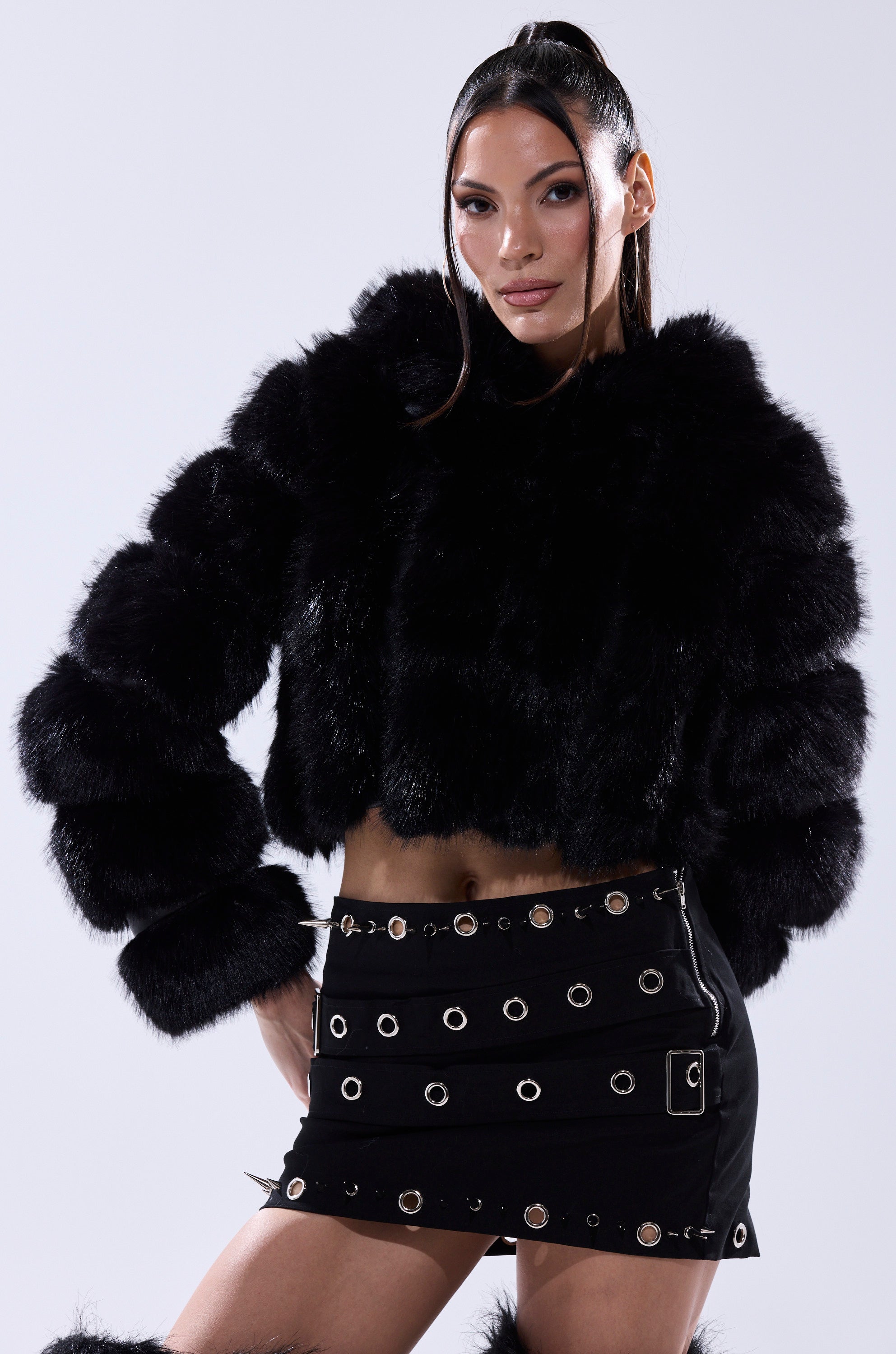 A woman poses confidently in the JOLENE FAUX FUR COAT IN BLACK and a black mini skirt with metallic grommet details, set against a light gray background.