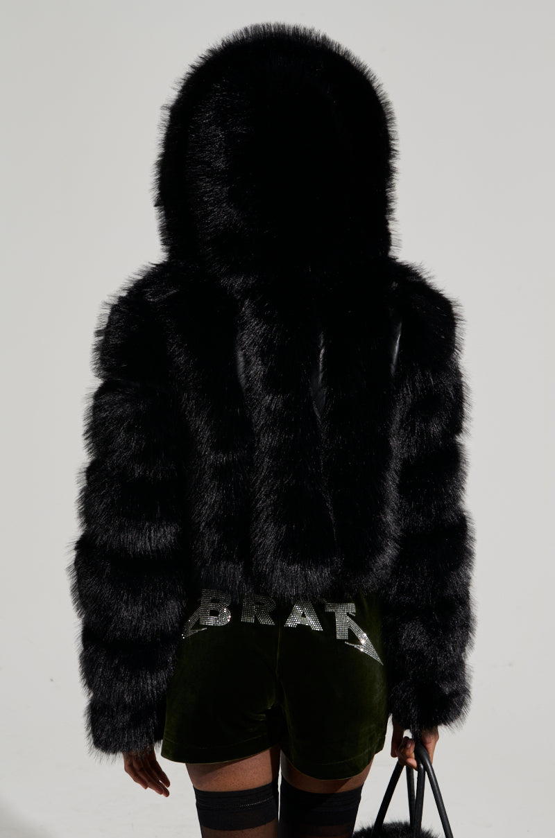 A person wears a stylish JOLENE FAUX FUR COAT IN BLACK with green velvet shorts adorned with "BRAT" in rhinestones. They are holding a black handbag and facing away from the camera.