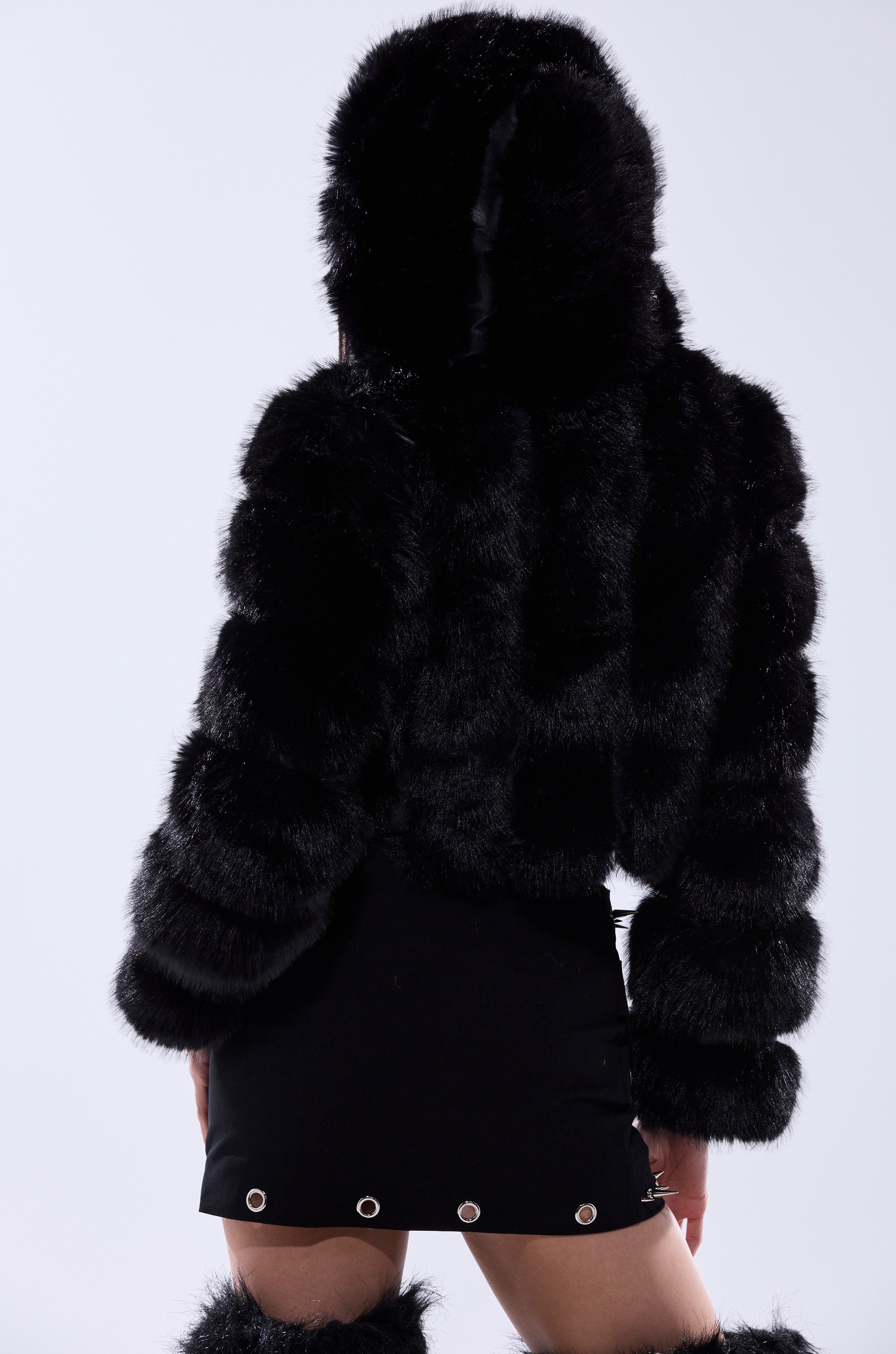 A person stands with their back to the camera against a plain light background, wearing the JOLENE FAUX FUR COAT IN BLACK with a sherpa-lined hood, paired with a black skirt featuring silver eyelets and fur-trimmed boots.