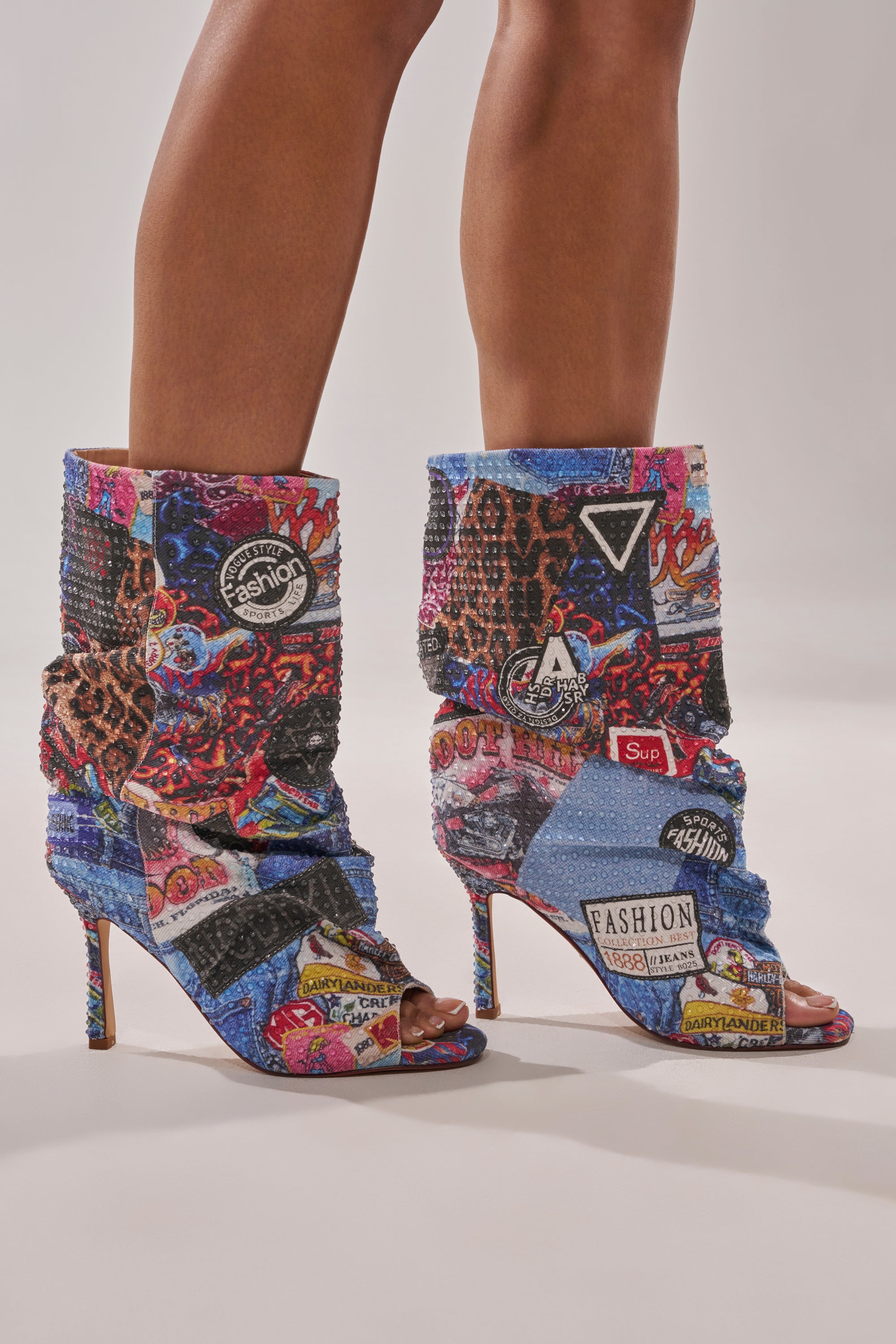 Someone stands confidently against a plain backdrop, wearing the AZALEA WANG JONICE BLUE MULTI RHINESTONE OPEN TOE BOOTIE, featuring high heels, open toes, and colorful rhinestone accents. 1