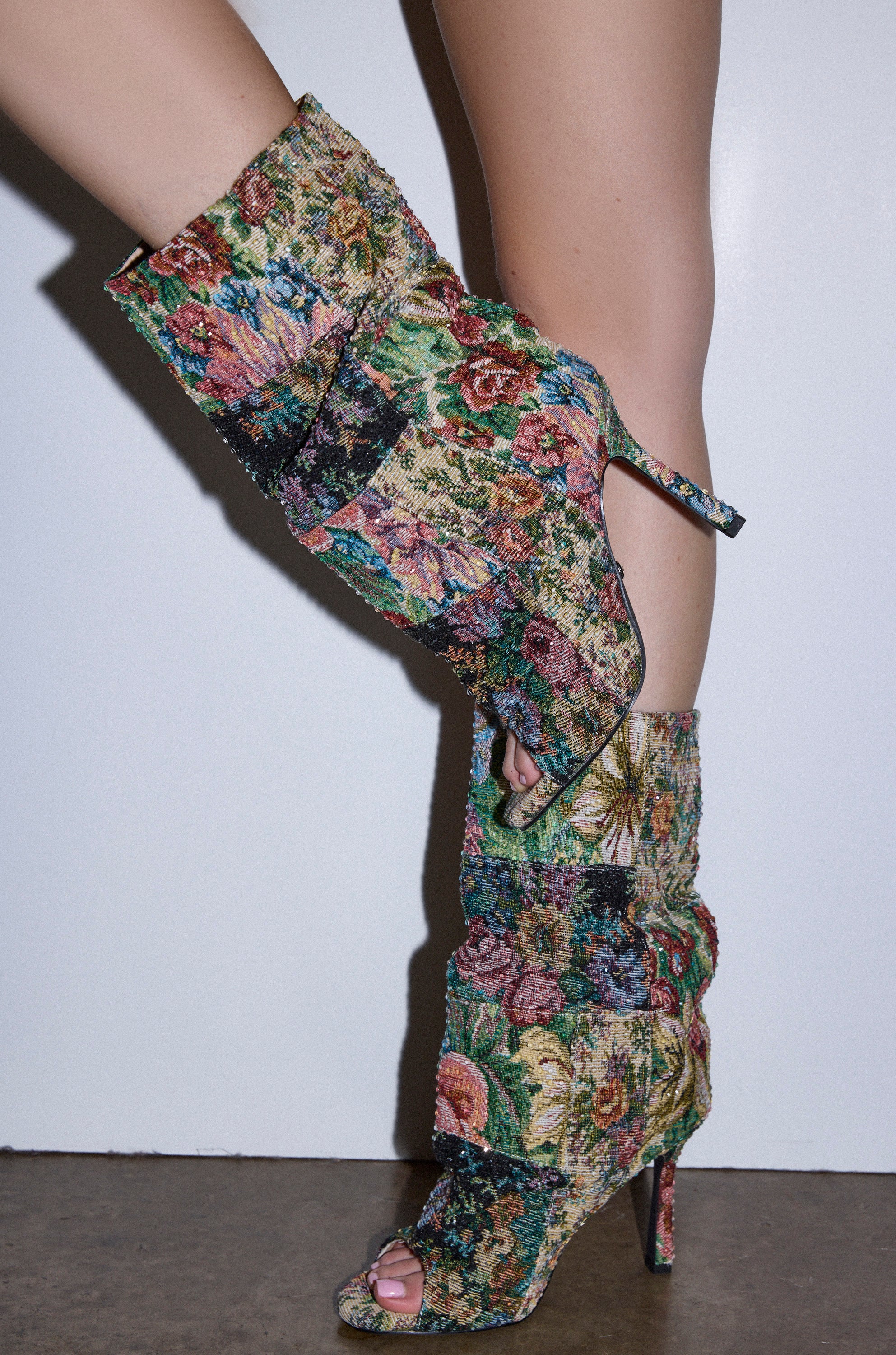 A close-up of legs wearing the AZALEA WANG JONICE FLORAL RHINESTONE EMBELLISHED OPEN TOE BOOTIE, featuring a patchwork tapestry design and stiletto heels against a plain white background.