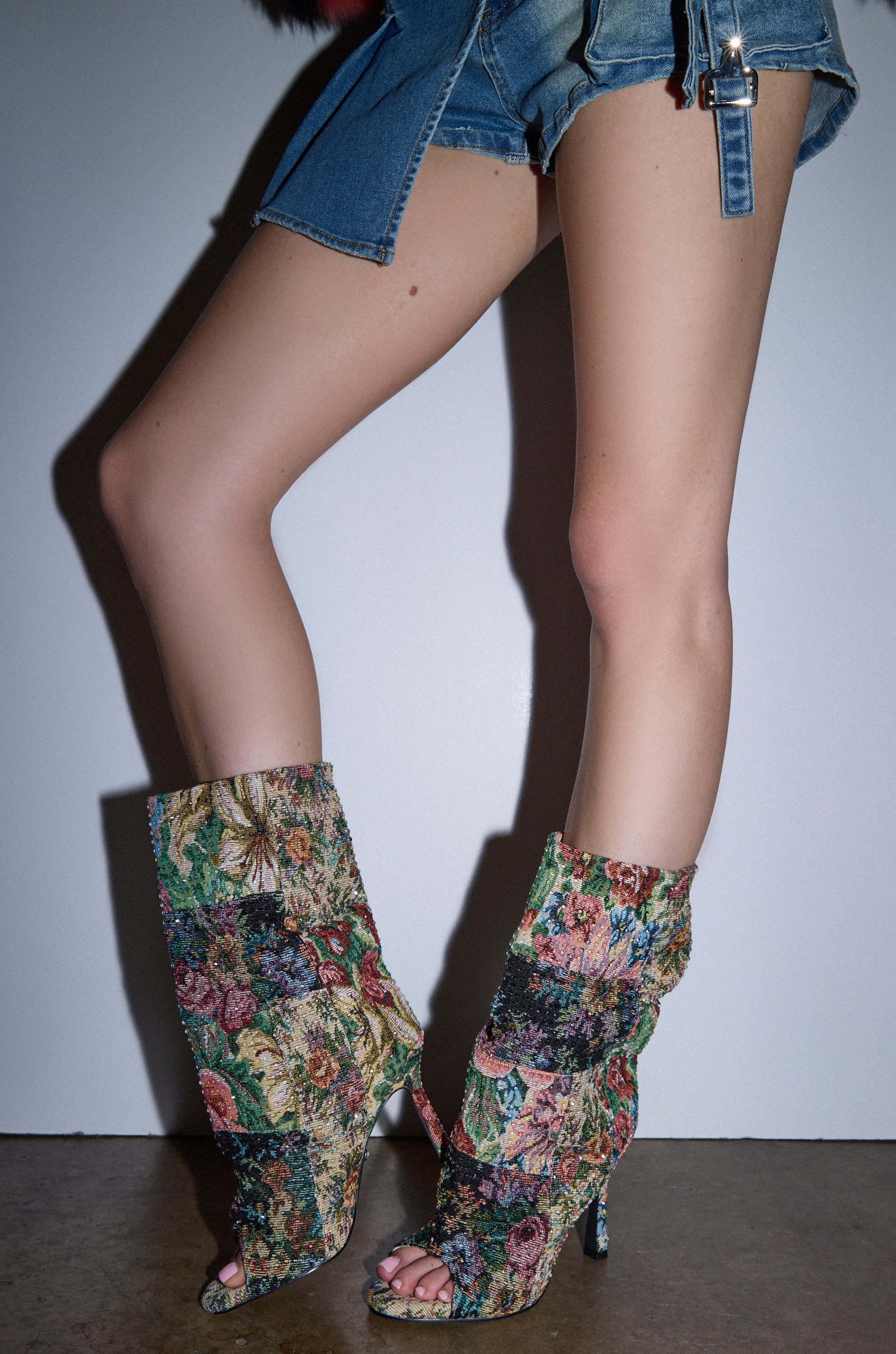 A person poses against a white wall, legs crossed at the ankles, wearing ripped denim shorts and AZALEA WANG JONICE Floral Rhinestone Embellished Open Toe Booties.