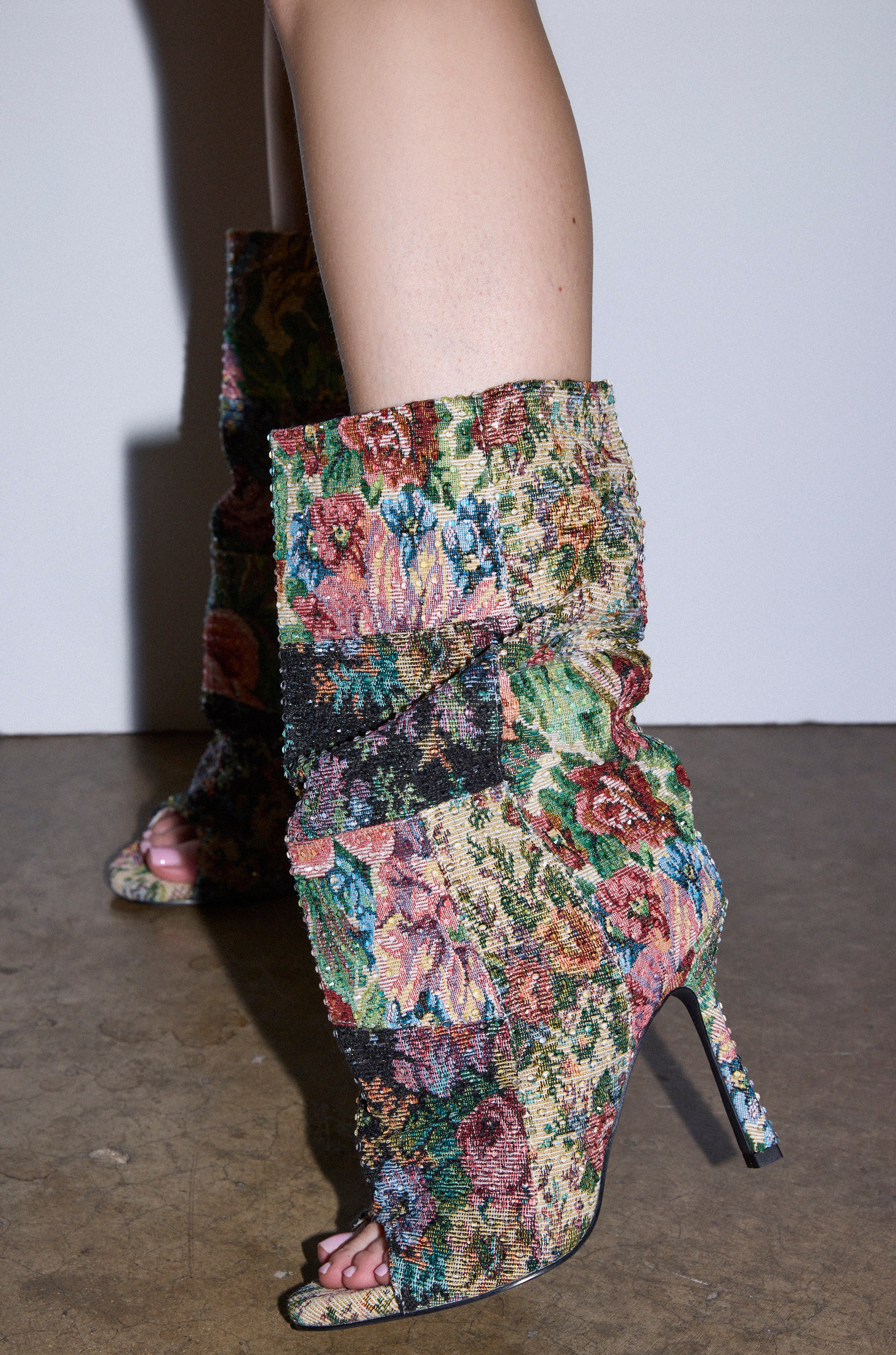 A person wears the AZALEA WANG JONICE FLORAL RHINESTONE EMBELLISHED OPEN TOE BOOTIE while standing on a concrete floor against a plain white wall.