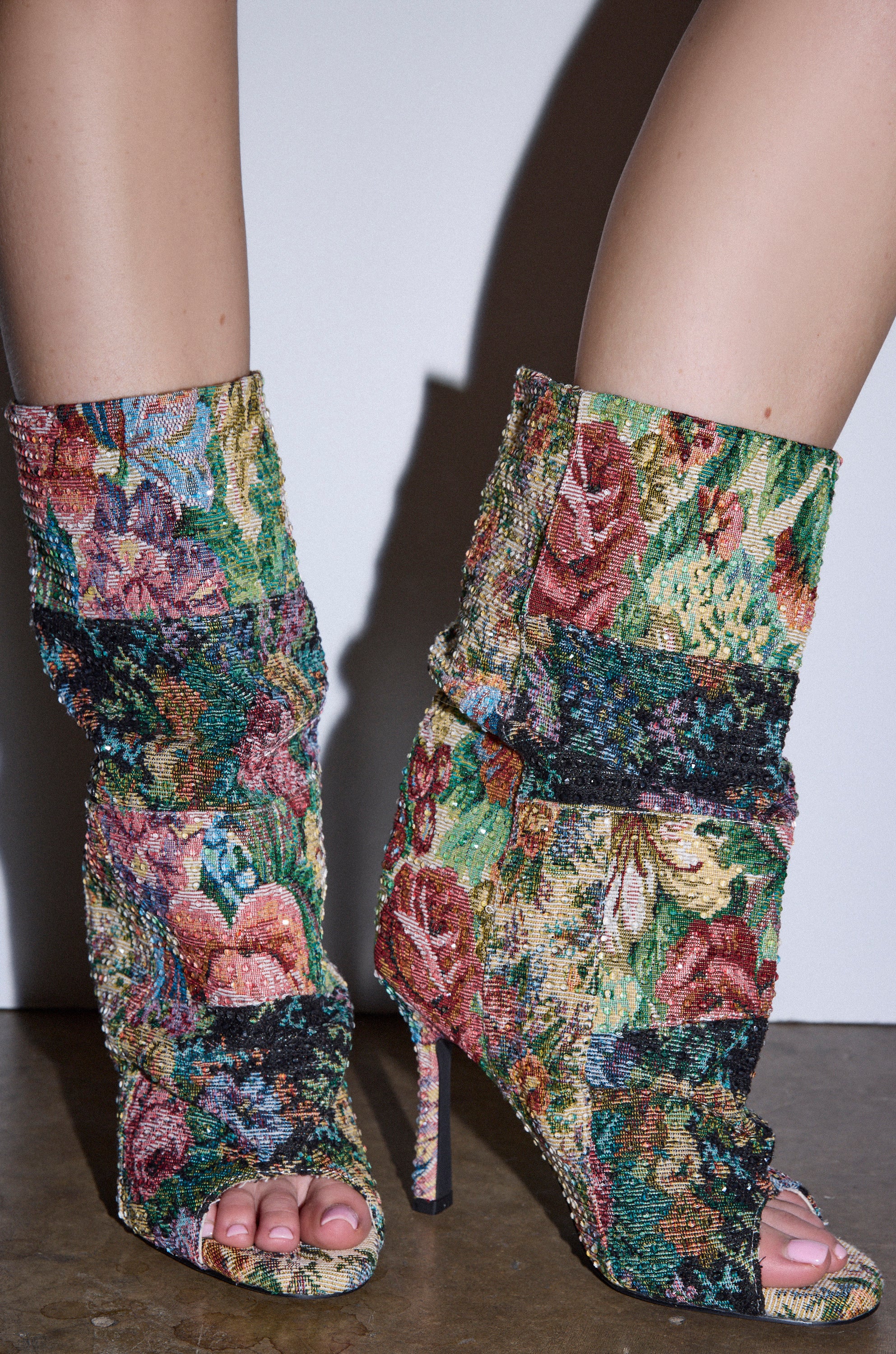 A close-up of two feet wearing the AZALEA WANG JONICE FLORAL RHINESTONE EMBELLISHED OPEN TOE BOOTIE, featuring a vibrant floral design, standing on a hard surface against a white background.