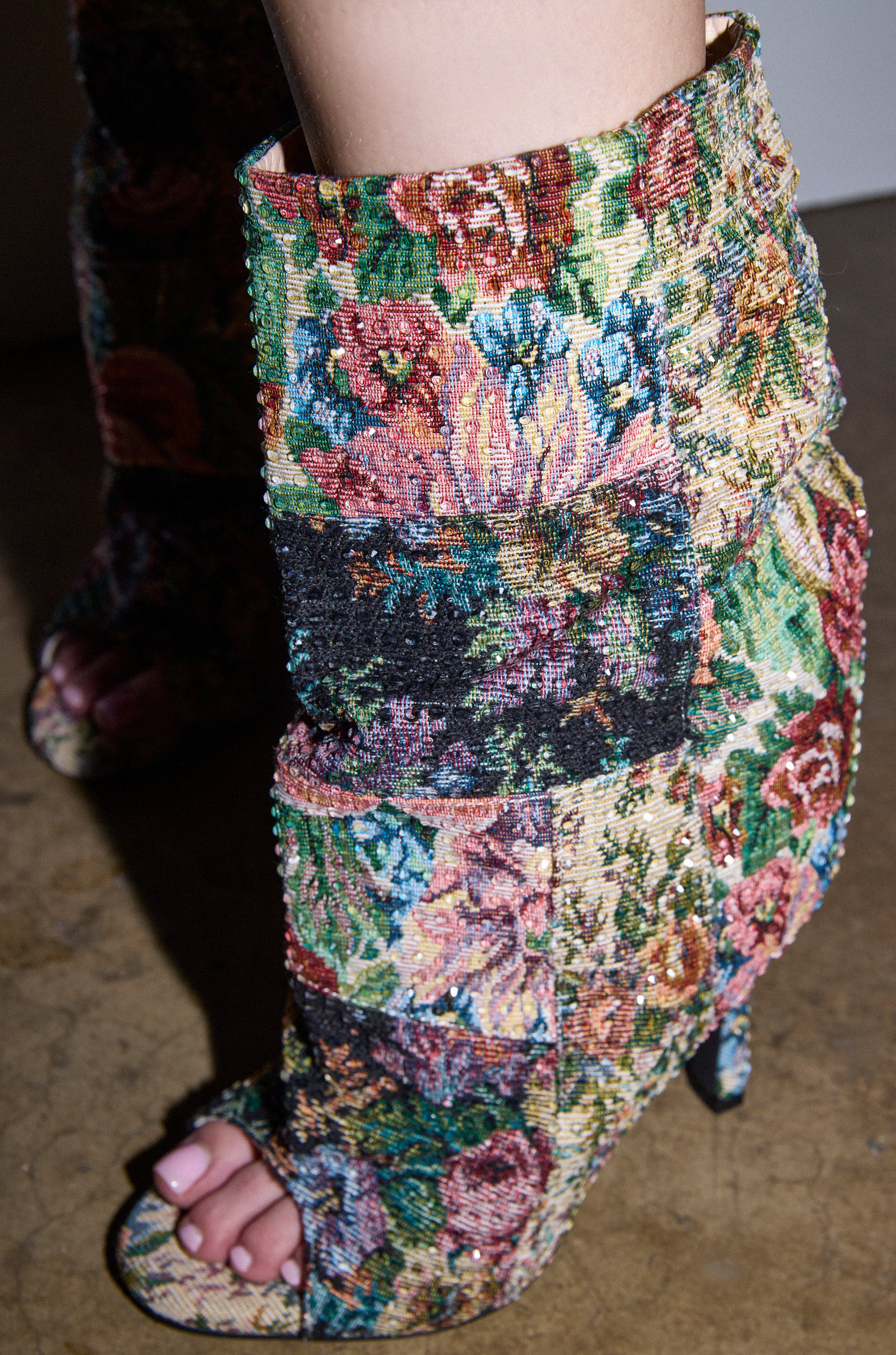 A close-up of a light-skinned foot with manicured toenails in the AZALEA WANG JONICE Floral Rhinestone Embellished Open Toe Bootie, stiletto heel, on a brown textured floor that highlights its vibrant design.
