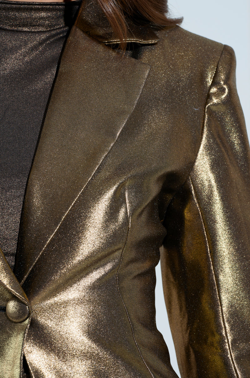 Close-up of the JOSH'S GOLD MEDAL JACKET from the AKIRA Label, worn over a dark turtleneck. The shimmering metallic gold fabric catches the light, highlighting its glossy texture and tailored fit. The image showcases part of the garment's lapel and a single button.