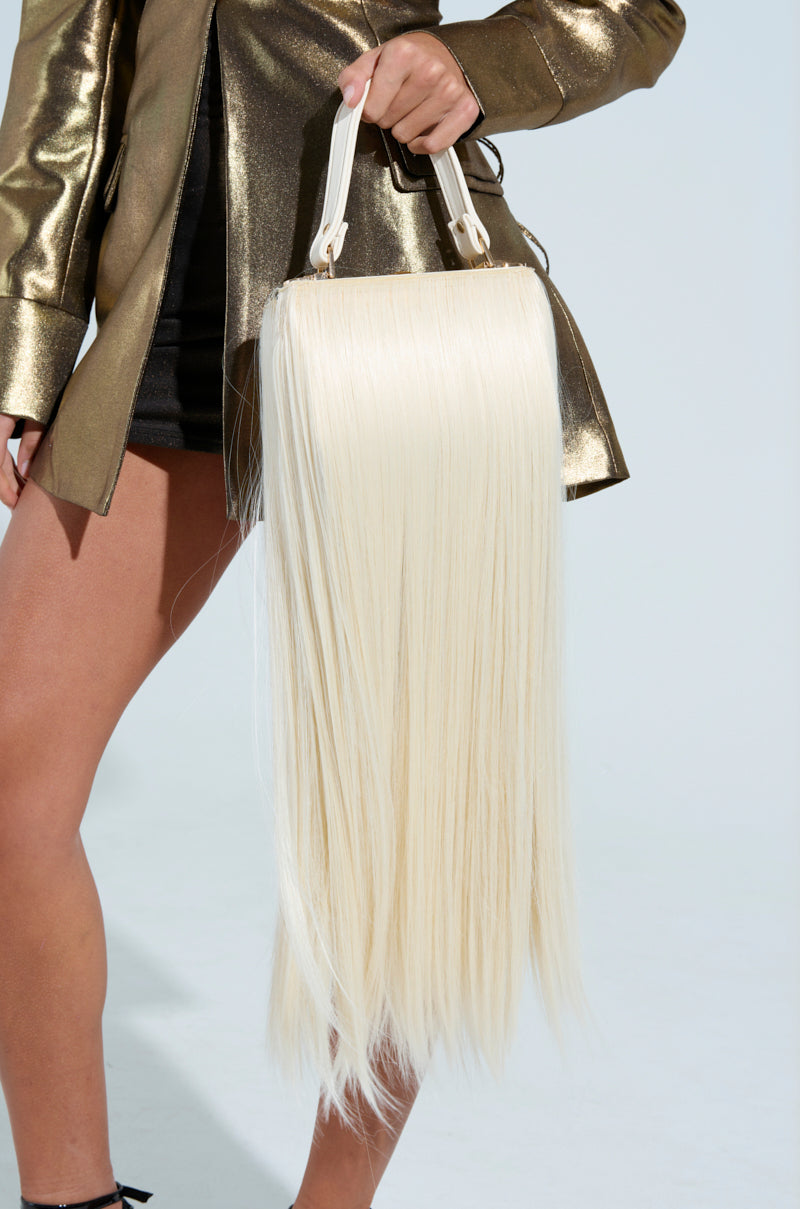 A person in a metallic gold jacket and black skirt holds a striking JOSHIE SYNTHETIC HAIR TRUNK PURSE adorned with long, straight, platinum blonde strands, set against a plain background.