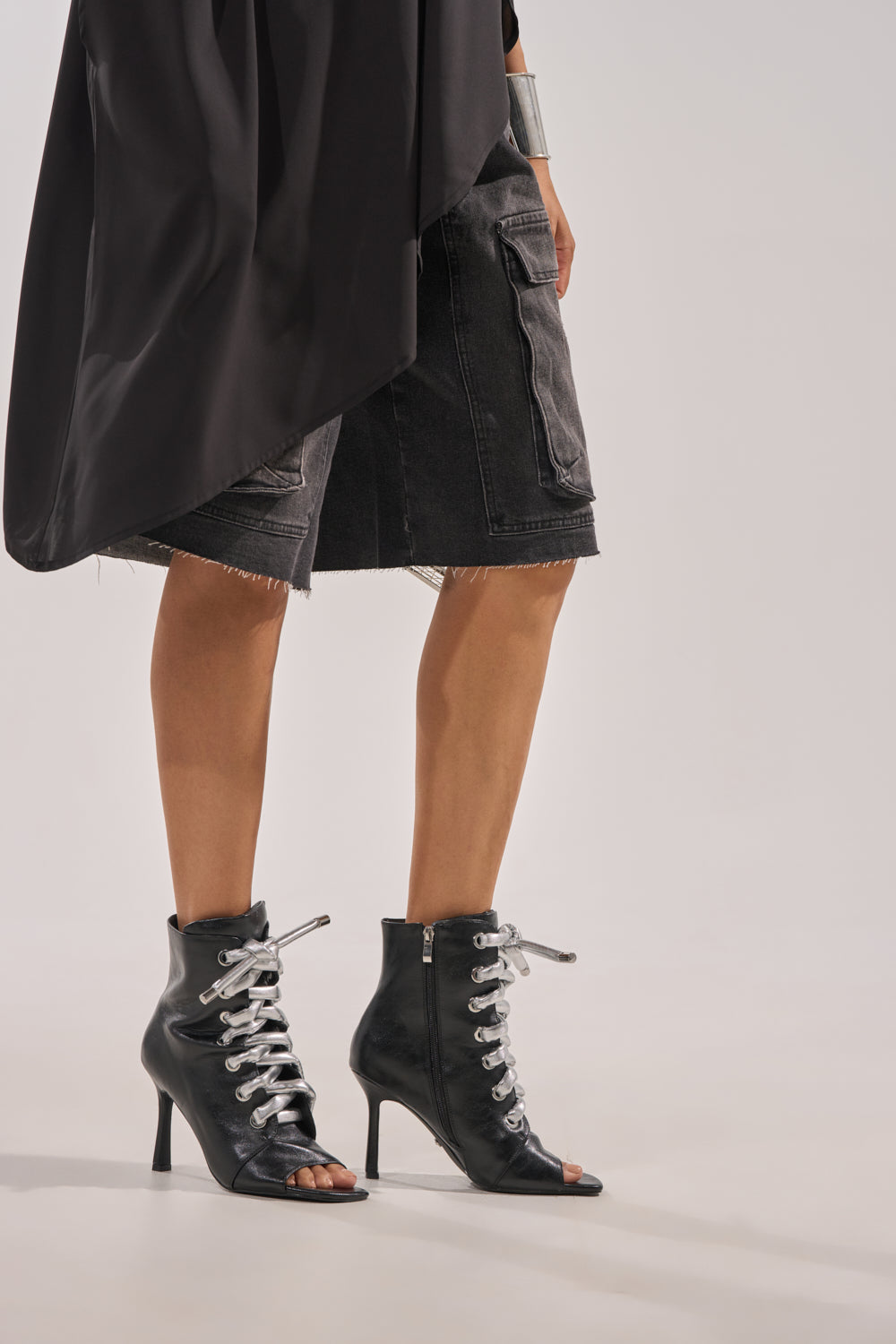 A person wears AZALEA WANG JOSS BLACK BLOCK LACE OPEN TOE BOOTIE, black denim knee-length shorts, and a black asymmetrical top. Only their lower body is visible against a plain light background. 1