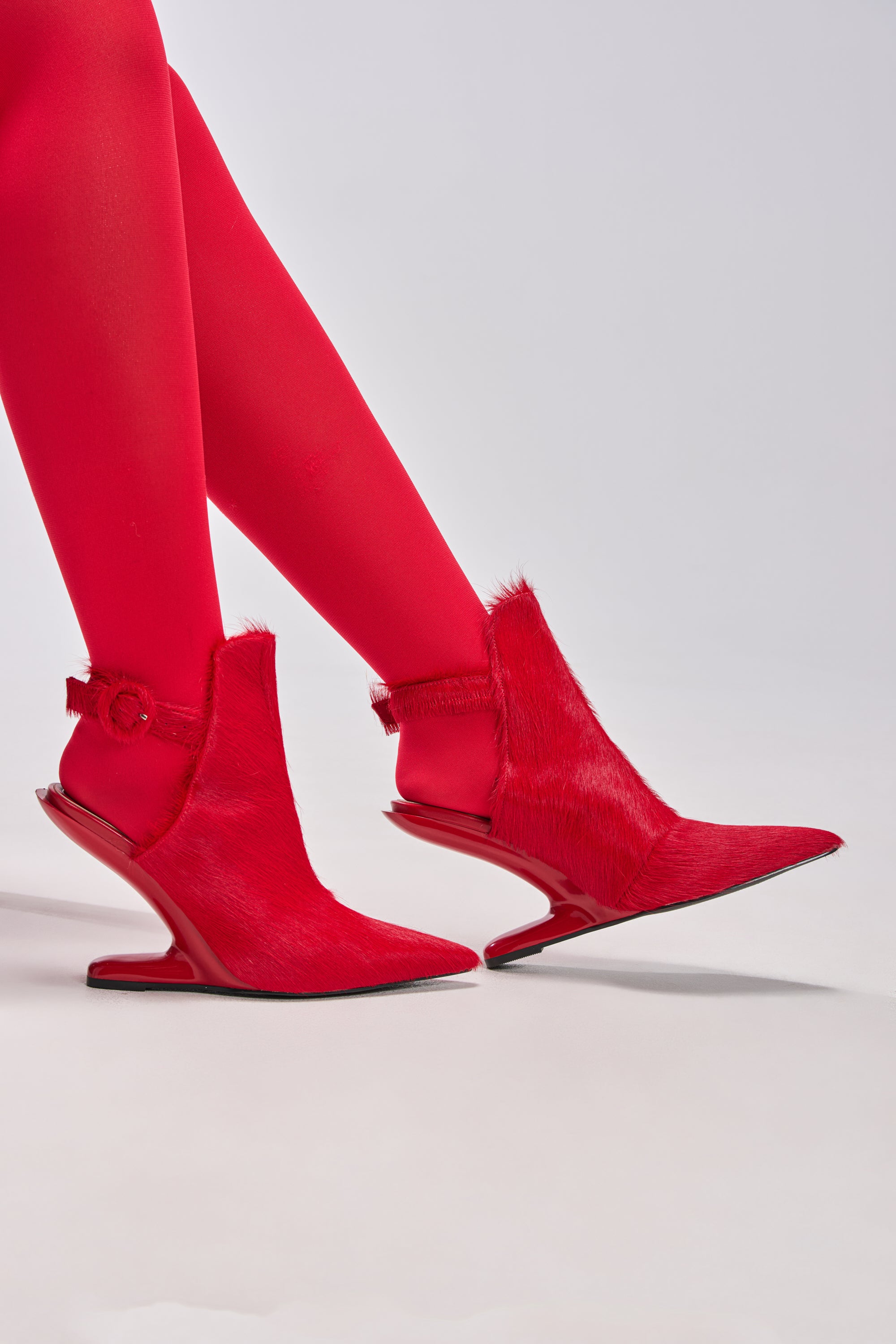 Wearing AZALEA WANG JOST RED PONY HAIR UPPER WEDGE PUMP, a person in bright red tights stands against a plain light background, showing off the pointed toes, furry pony hair texture, and angled red wedge heels of the unique shoes. 2