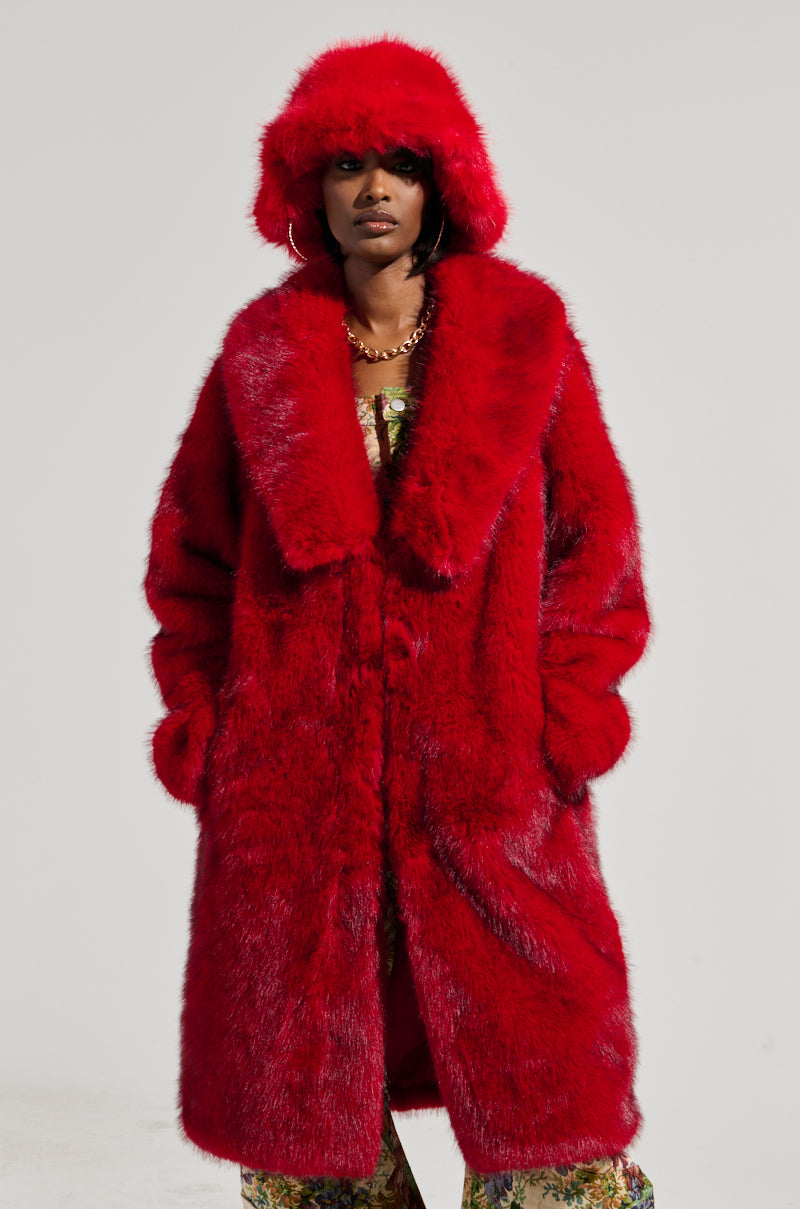 Dressed in the JOVI FAUX FUR JACKET, a person stands confidently with their hands in their coat pockets. They are adorned in a vibrant red faux fur jacket paired with a matching fur hat and oversized floral patterned pants. The background is a simple, light gray.