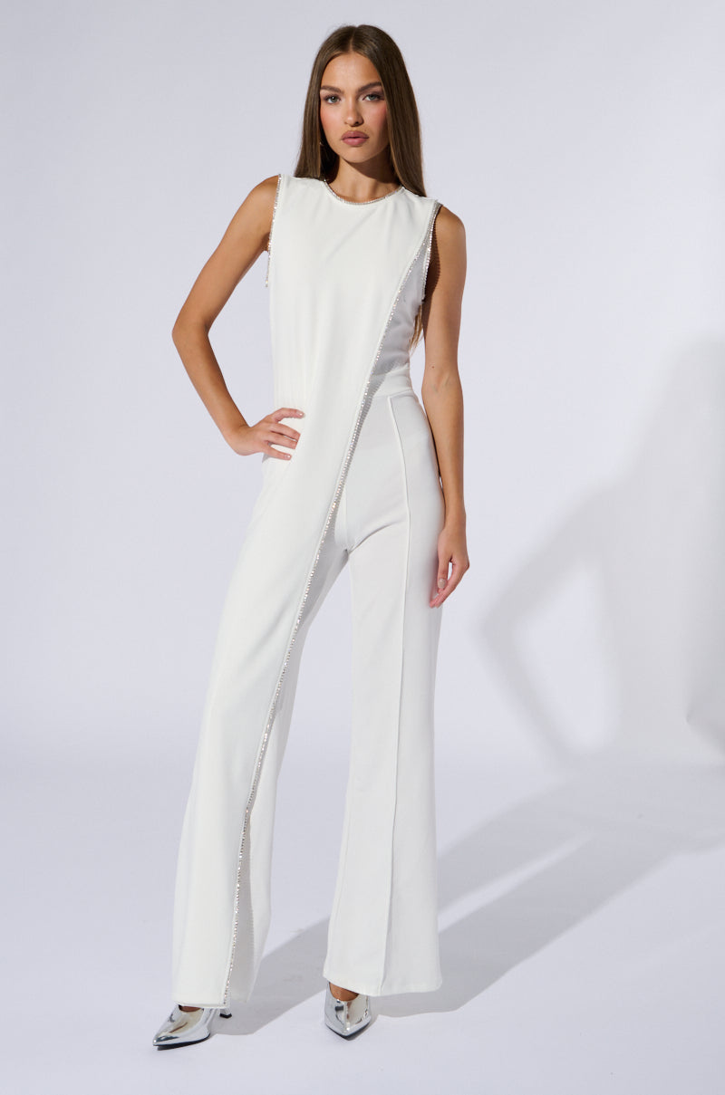 A woman stands in a three-quarter pose against a plain white background, wearing the NOTHING BUT CLASS WIDE LEG JUMPSUIT featuring an asymmetrical design with a tonal diagonal fabric overlay. She has long, straight hair and is wearing silver pointed-toe heels adorned with rhinestone chain link trim. She gazes confidently at the camera.