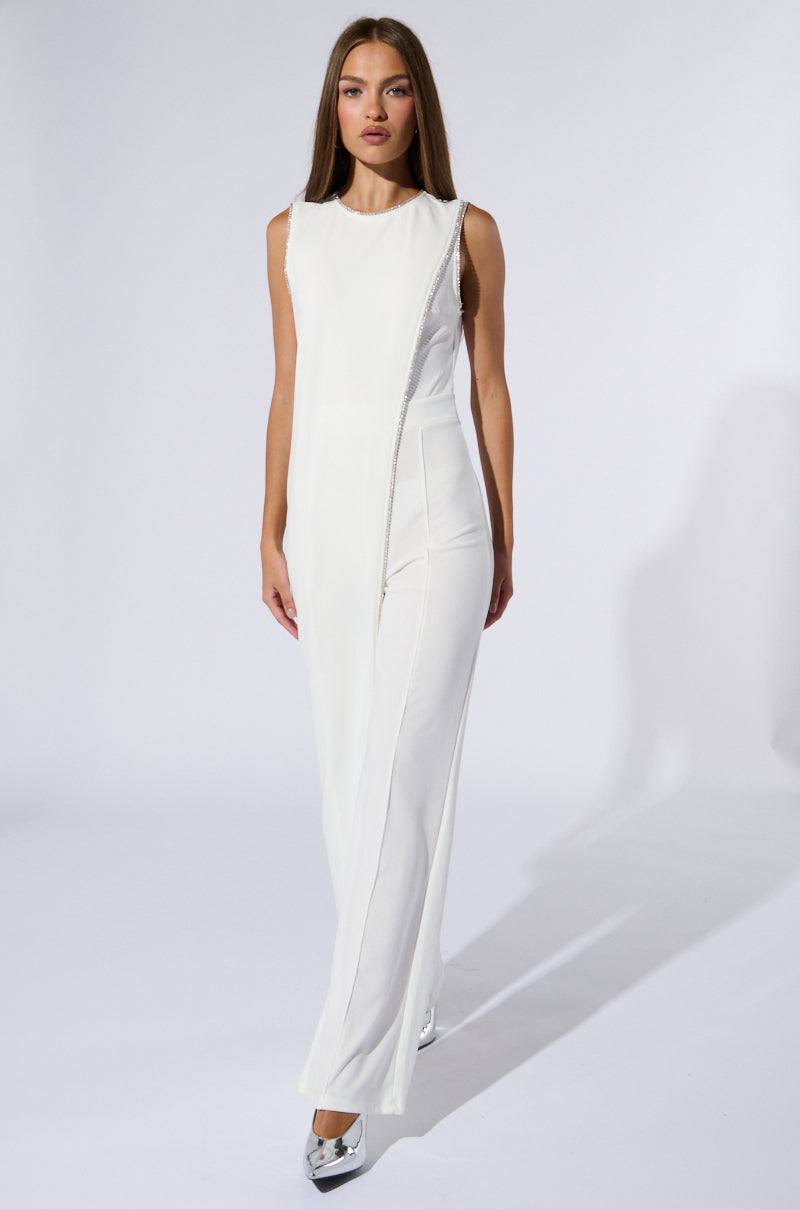 A woman with long hair walks confidently towards the camera, dressed in the NOTHING BUT CLASS WIDE LEG JUMPSUIT, a sleek, sleeveless white outfit featuring a tonal diagonal fabric overlay. She is wearing metallic silver high-heeled shoes and is set against a plain white background.