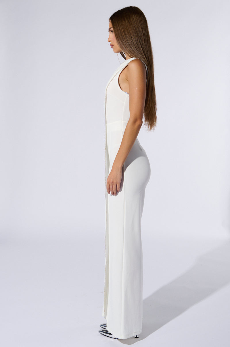 A woman with long, straight hair wearing the "Nothing But Class Wide Leg Jumpsuit," a white, sleeveless outfit highlighted by a rhinestone chain link trim, and black-and-white striped shoes stands in profile against a plain white background. She gazes forward, her arms relaxed by her sides.