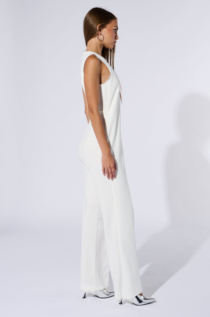 A person standing in profile is wearing the NOTHING BUT CLASS WIDE LEG JUMPSUIT, featuring a sleeveless, white design. They also have on black-and-white shoes and large, circular earrings. The plain white background highlights their straight, long hair that falls down their back, beautifully accentuating the jumpsuit's tonal diagonal fabric overlay.