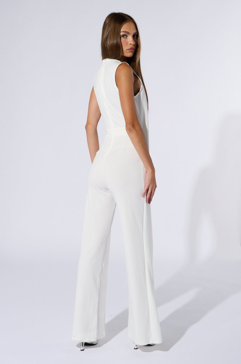 A woman with long, straight brown hair is standing and turned slightly to her left, looking over her shoulder. She is wearing the NOTHING BUT CLASS WIDE LEG JUMPSUIT, a sleeveless white jumpsuit with a tonal diagonal fabric overlay, paired with high-heeled shoes. The background is plain and white.