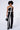 A woman with short curly hair stands against a white background, dressed in the LIONA JUMPSUIT. The outfit showcases a strapless design and wide leg silhouette, accentuated by bold white diagonal and vertical stripes. She carries a black handbag and wears black pointed-toe heels.