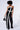 A woman wearing the Liona Jumpsuit, a strapless black jumpsuit with striking white diagonal and vertical stripes and a wide leg silhouette. She holds a black handbag and has short curly hair, looking down to her left side against a plain white background.