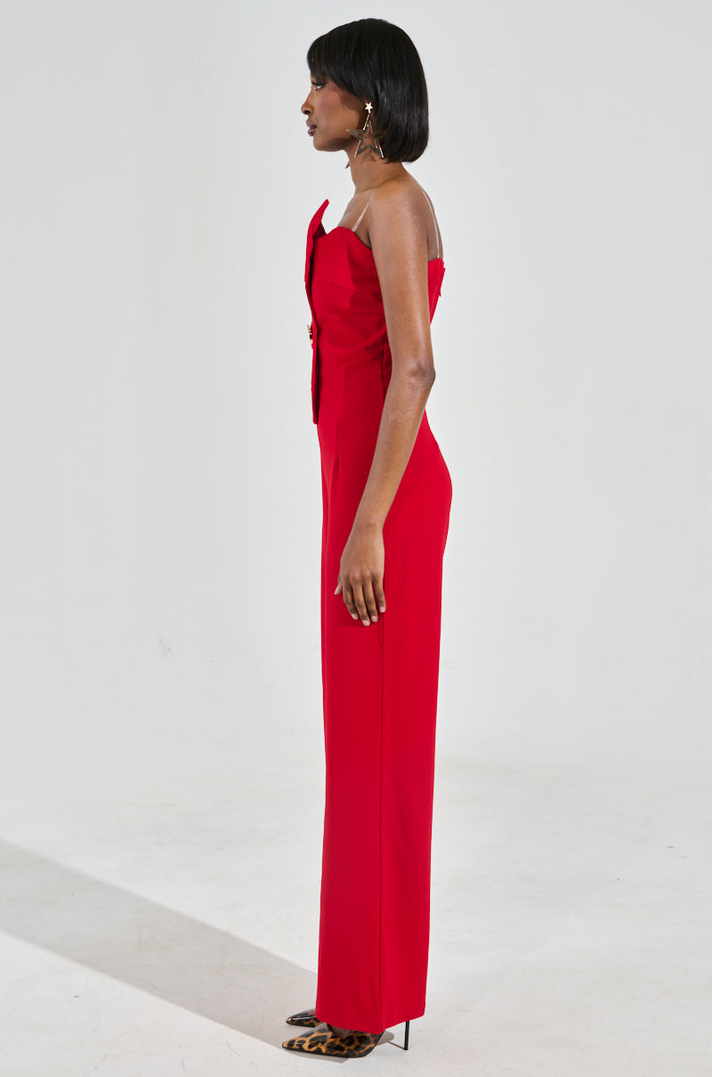 A person in profile stands against a light gray background, showcasing a BADDIE STRAPLESS JUMPSUIT WITH GOLD BUTTONS in vivid red, paired with leopard-print high heels. The chic jumpsuit features a bow detail on the front, while their dark hair is styled straight, exuding confidence and flair.