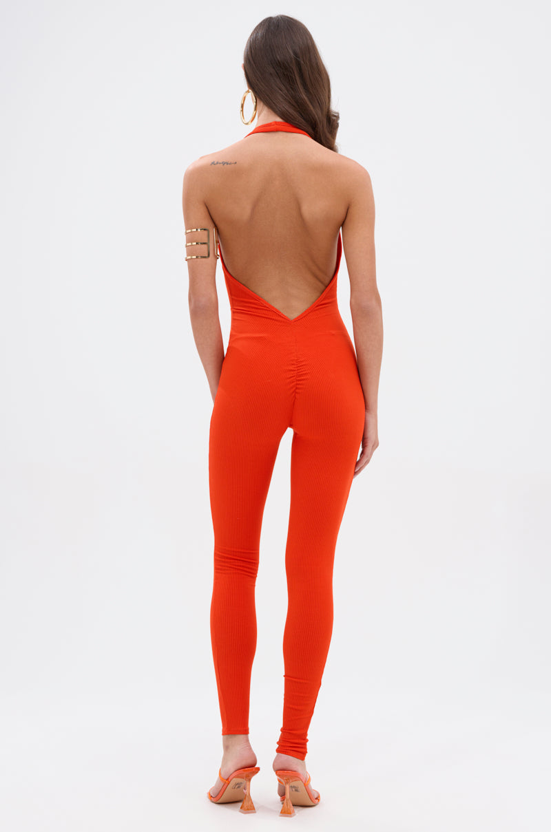 A woman, facing away against a white background, models the KEEP IT CASUAL HALTER JUMPSUIT IN ORANGE with orange heeled sandals, large hoop earrings, an arm cuff, and flowing long brown hair for a chic look.