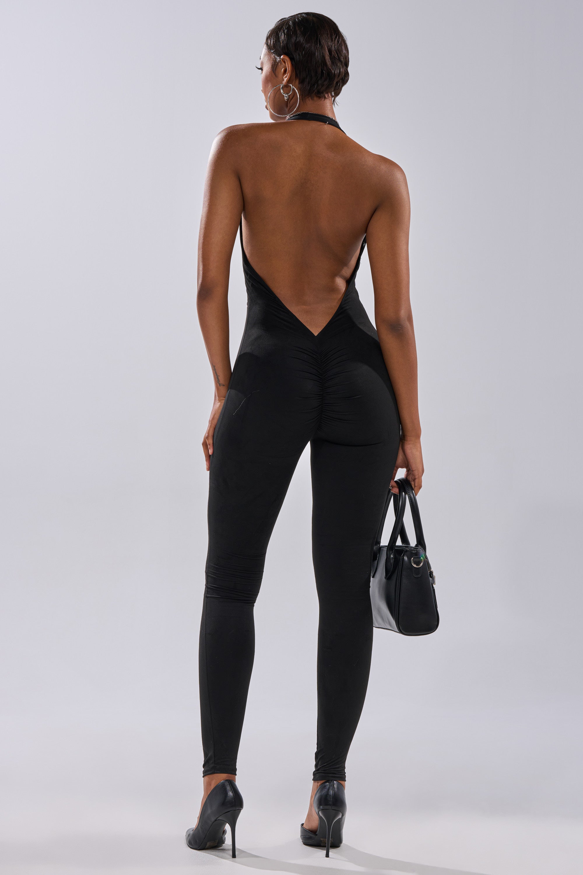 A person wearing the GIA SUEDE JUMPSUIT, featuring a black, curve-hugging, halter style, stands facing away with high heels, a black handbag, and hoop earrings against a plain light background.
