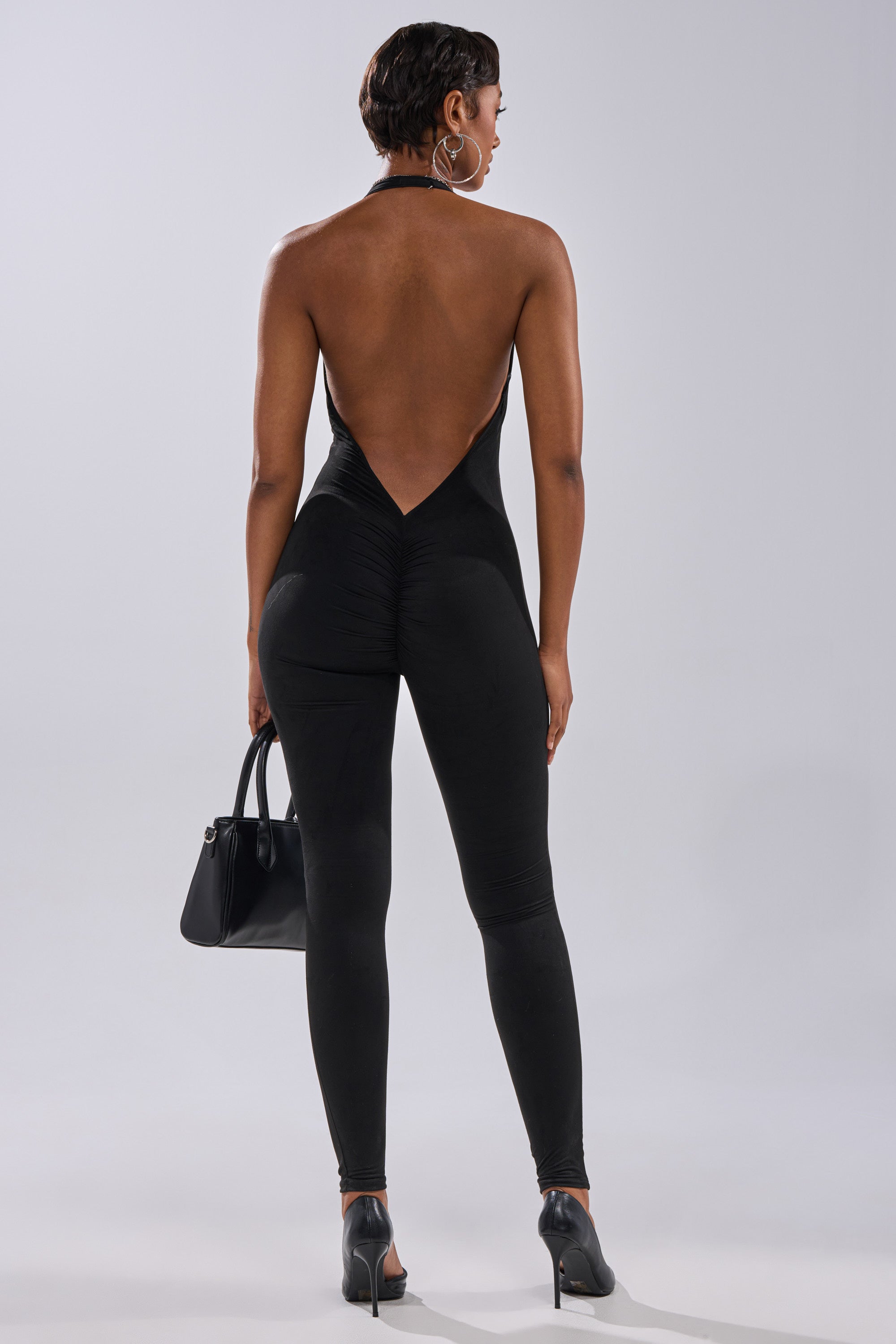 A woman, her short hair styled with large hoop earrings, faces away wearing the GIA SUEDE JUMPSUIT featuring a halter V-neck and deep V-cut back. She pairs it with black heels and carries a small black handbag.