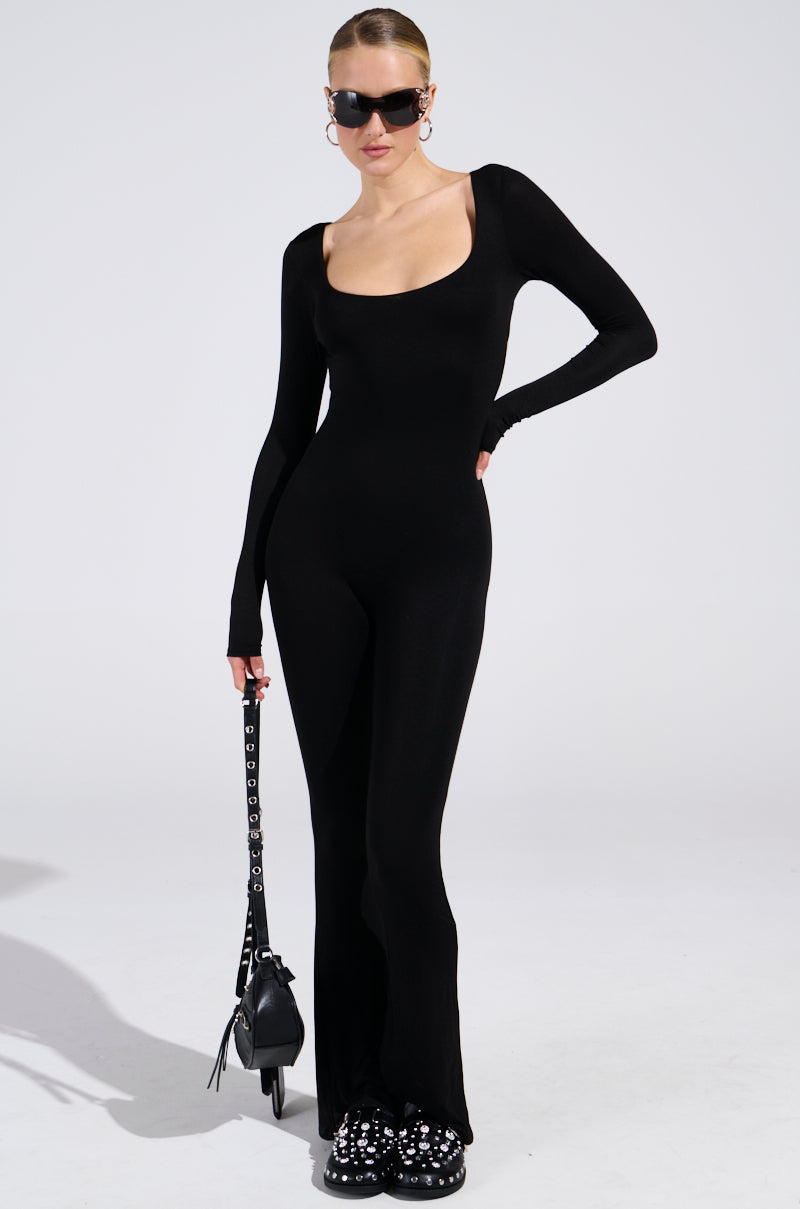 Model in the "IN YOUR DREAMS JUMPSUIT IN BLACK" poses confidently. Wearing sunglasses and large hoop earrings, they hold a black bag with a studded strap. Their hair is styled back, and statement shoes complete the look against a plain white background.