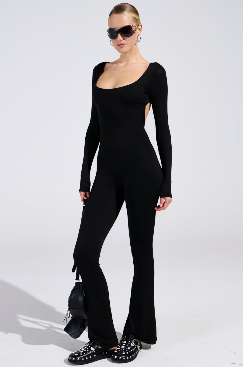 Dressed in the "IN YOUR DREAMS JUMPSUIT IN BLACK," a person stands against a plain backdrop, enhanced by sunglasses and studded black shoes. They hold a small black handbag, with their hair pulled back, showcasing a curve-accentuating silhouette.