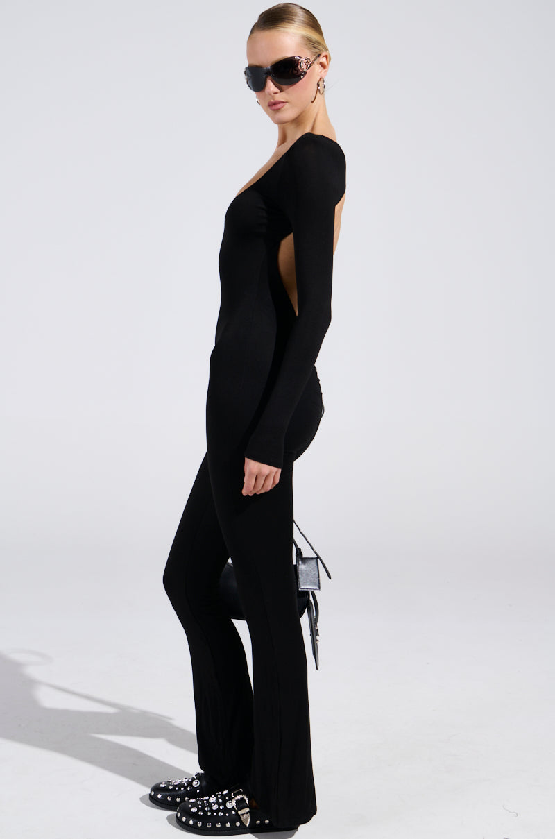 Wearing the IN YOUR DREAMS JUMPSUIT IN BLACK, a person shows off a curve-accentuating silhouette with an open-back design. Large sunglasses, studded shoes, and a small handbag elegantly adorn the ensemble against a neutral background.