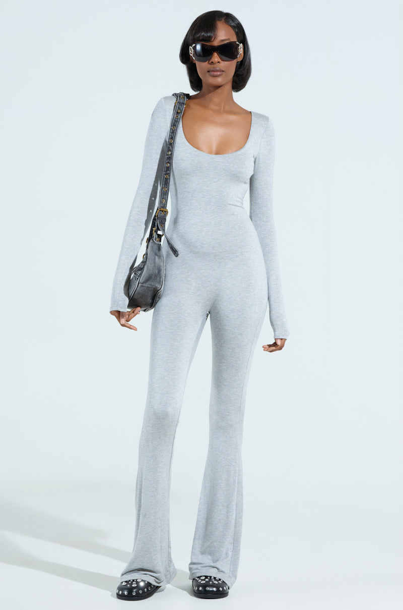 A woman confidently stands on a light background wearing the IN YOUR DREAMS JUMPSUIT with long sleeves, paired with black platform shoes and large black sunglasses, holding a black shoulder bag.