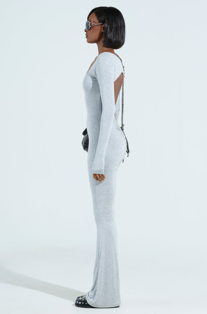 A person with a short bob haircut is wearing the IN YOUR DREAMS JUMPSUIT, a long-sleeved, curve-clinging light gray outfit with an open back. They have a black crossbody bag and sunglasses, standing in profile against a plain backdrop.