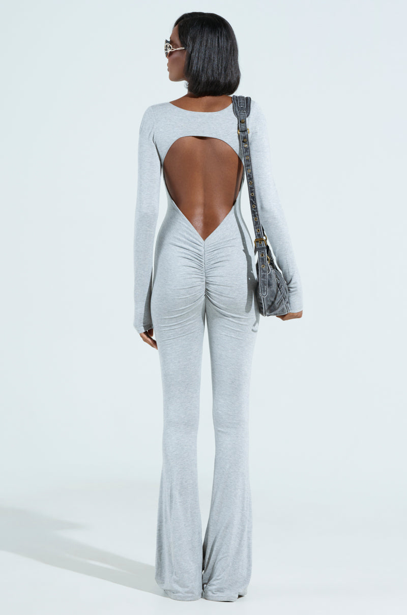 A woman with short black hair wears the IN YOUR DREAMS JUMPSUIT, a curve-hugging grey outfit with ruched detailing and flared legs, paired with a matching shoulder bag and sunglasses, against a pale blue background.