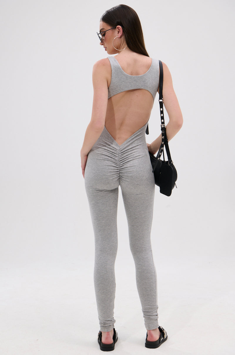 A woman with long straight hair and sunglasses faces away, wearing the gray IN YOUR DREAMS SLEEVELESS JUMPSUIT featuring a curve-hugging fit, open back, and ruched detailing. She pairs it with sandals and a black shoulder bag against a white background.