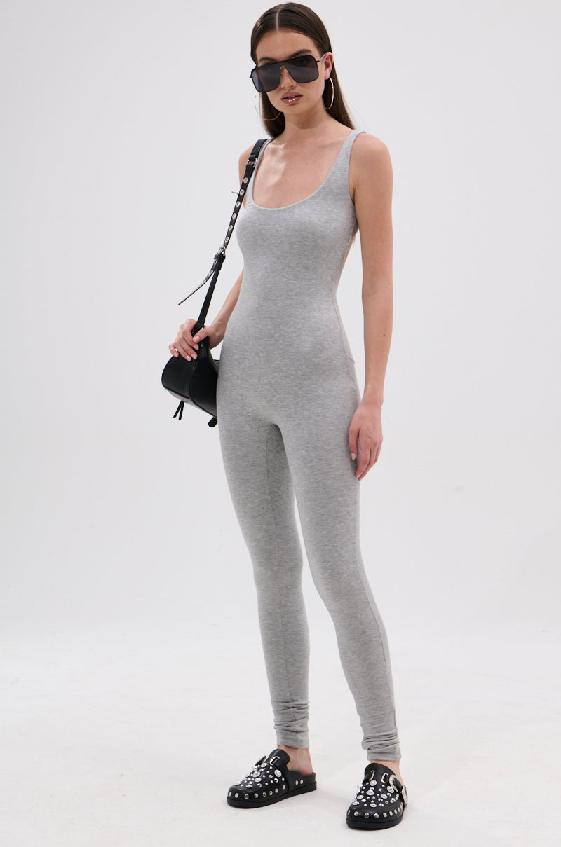 Woman in the IN YOUR DREAMS SLEEVELESS JUMPSUIT, a fitted light gray sleeveless piece made of stretchy knit fabric, styled with black studded slip-on shoes, a black shoulder bag, large hoop earrings, and dark sunglasses against a white background.