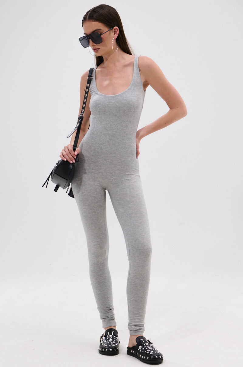 A woman models the IN YOUR DREAMS SLEEVELESS JUMPSUIT in light gray, featuring a fitted silhouette, paired with black studded shoes, large hoop earrings, oversized sunglasses, and a small black shoulder bag against a white background.