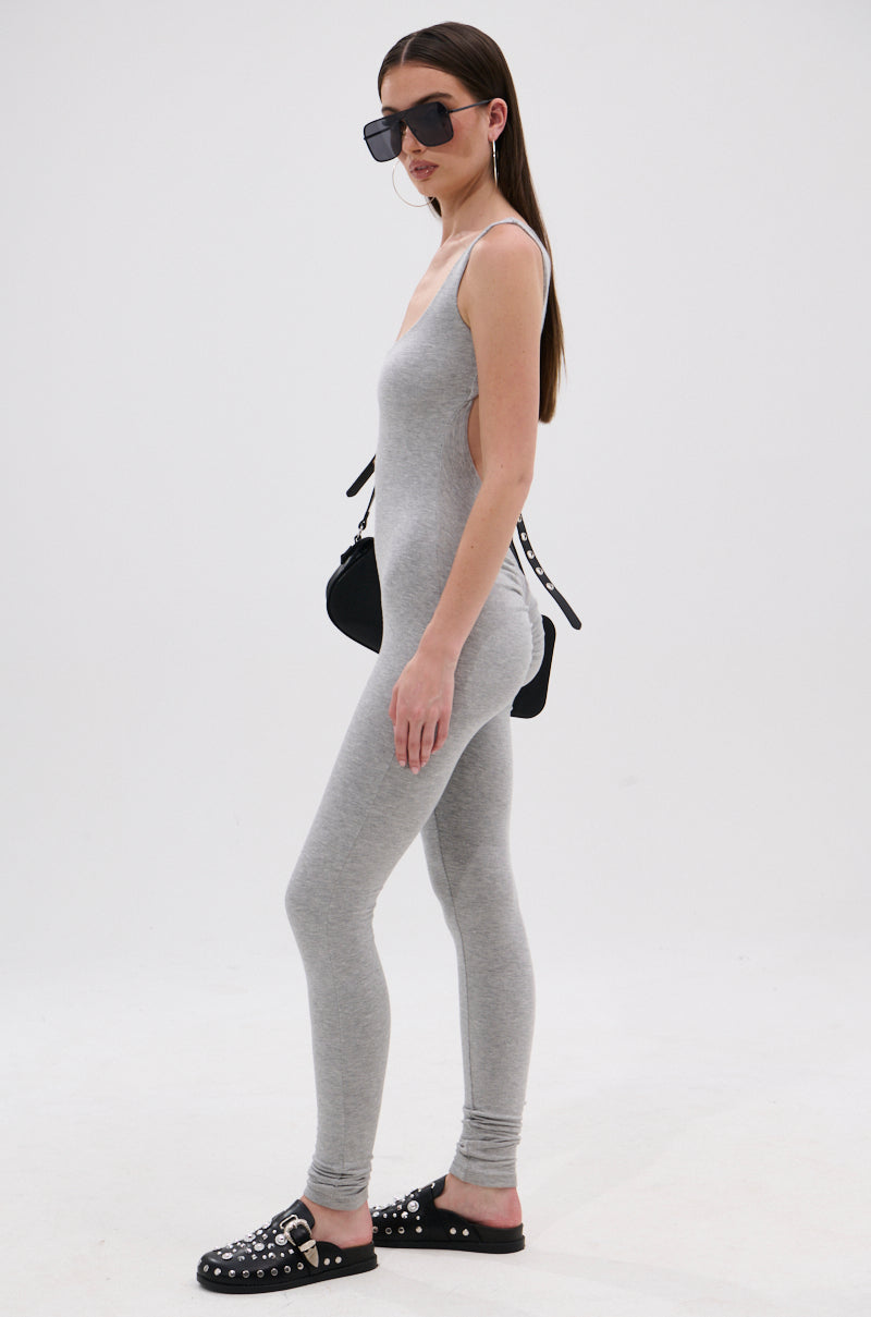 A woman models the IN YOUR DREAMS SLEEVELESS JUMPSUIT in grey, featuring a curve-hugging silhouette. She accessorizes with large black sunglasses, black studded slip-on shoes, and a black shoulder bag against a plain white background.