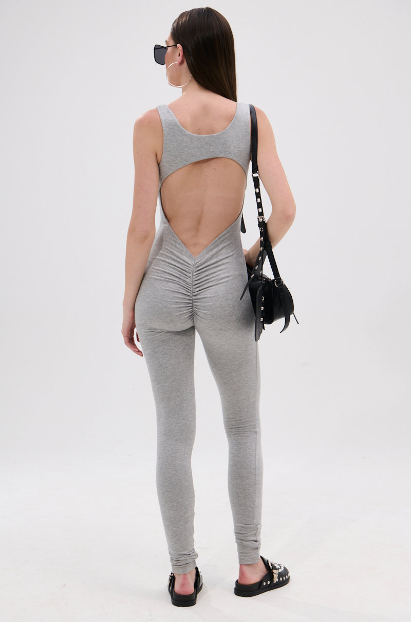 A woman with long straight hair and sunglasses faces away, wearing the IN YOUR DREAMS SLEEVELESS JUMPSUIT in light gray. The stretchy knit piece features an open back and ruched detailing. She accessorizes with a black bag and sandals against a white background.