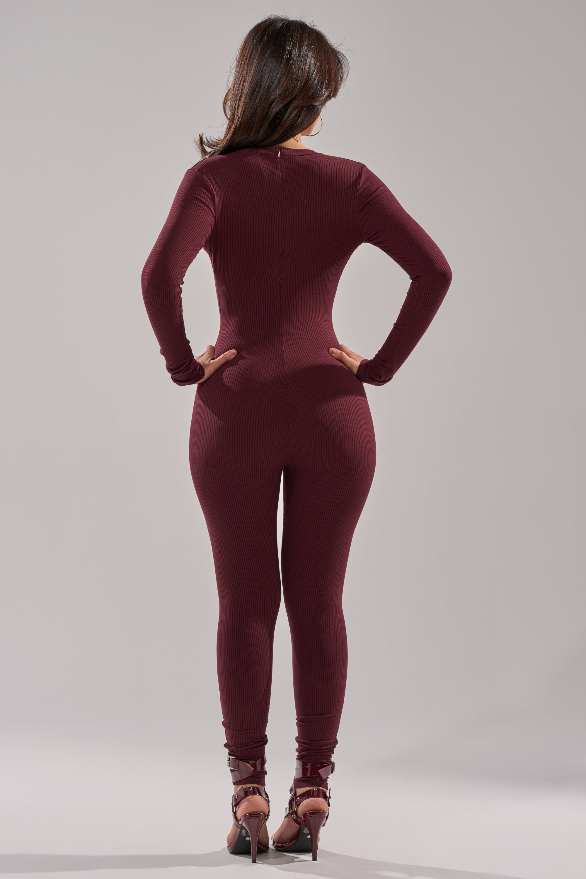 A woman faces away from the camera, hands on hips, wearing the DEEP V LONG SLEEVE JUMPSUIT IN RAISIN and high-heeled sandals against a plain light gray background. 2